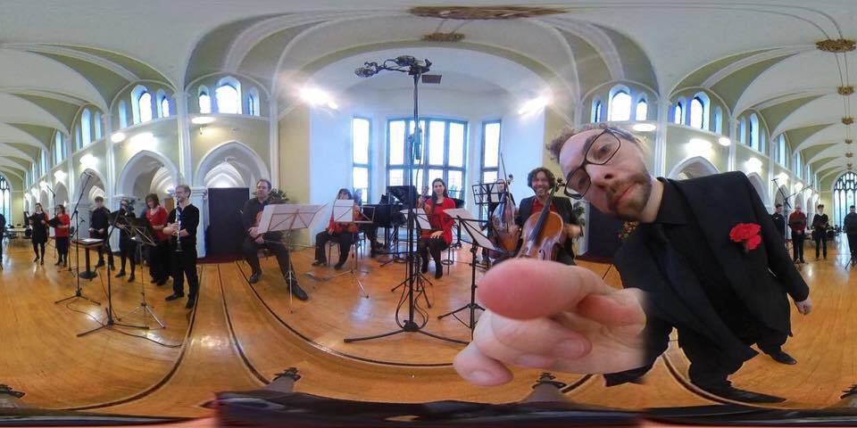 A group of musicians performing in a bright, arched hall with large windows, with a man in the foreground pointing at the camera.