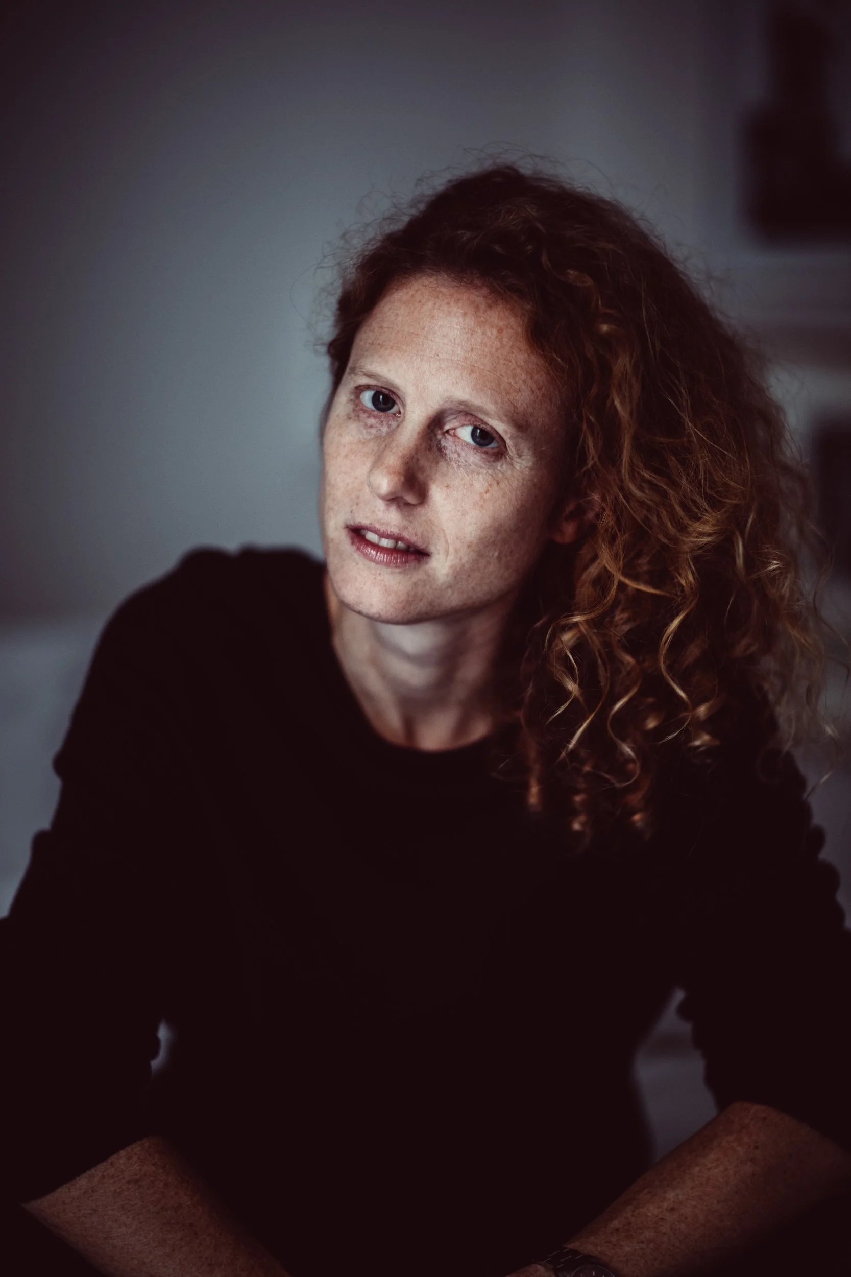 Close-up of a woman with curly red hair and freckles, wearing a black top, sitting at a table with her hands resting on it, in a dimly lit room.