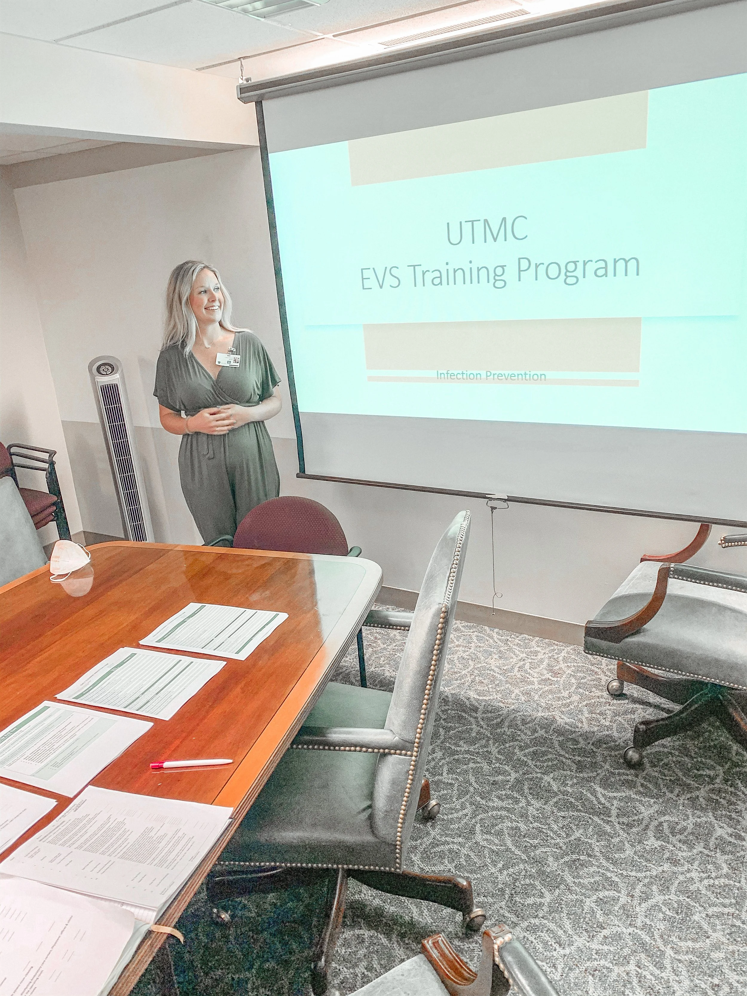 A woman standing in front of a projection screen that says "UTMC EVS Training Program" with the subtitle "Infection Prevention." She is smiling, wearing a dark green dress, and has a name badge. The room has a wooden conference table with papers and a red pen, surrounded by chairs, and a large fan is visible in the background.