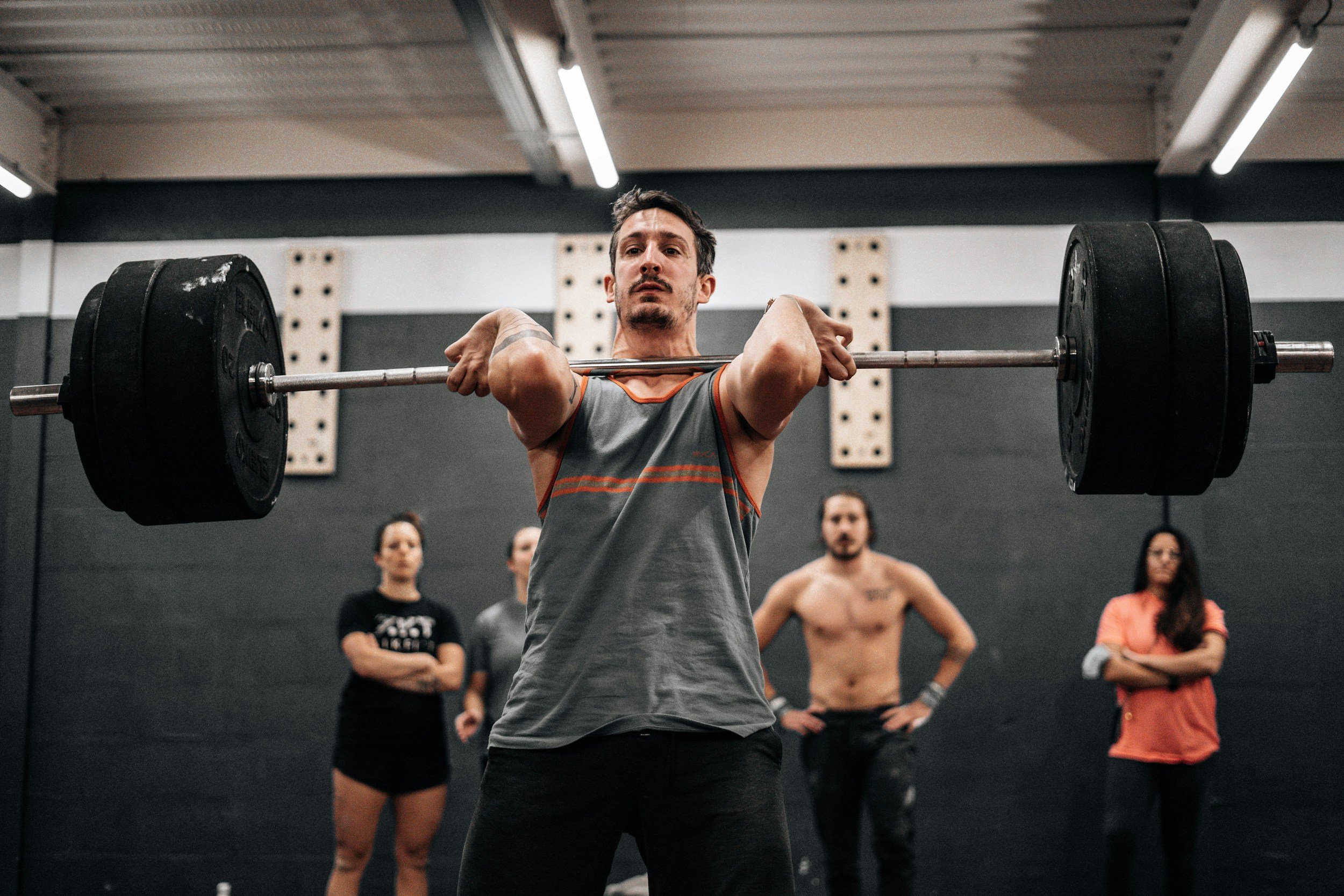 A man lifting a barbell with weights at a gym, surrounded by people watching.