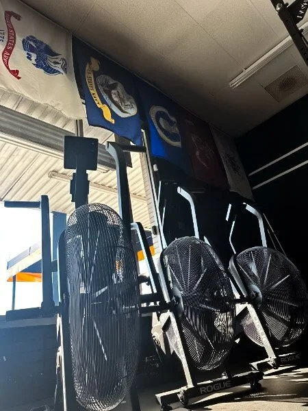 Indoor gym with flags hanging from the ceiling and large fans on exercise equipment.