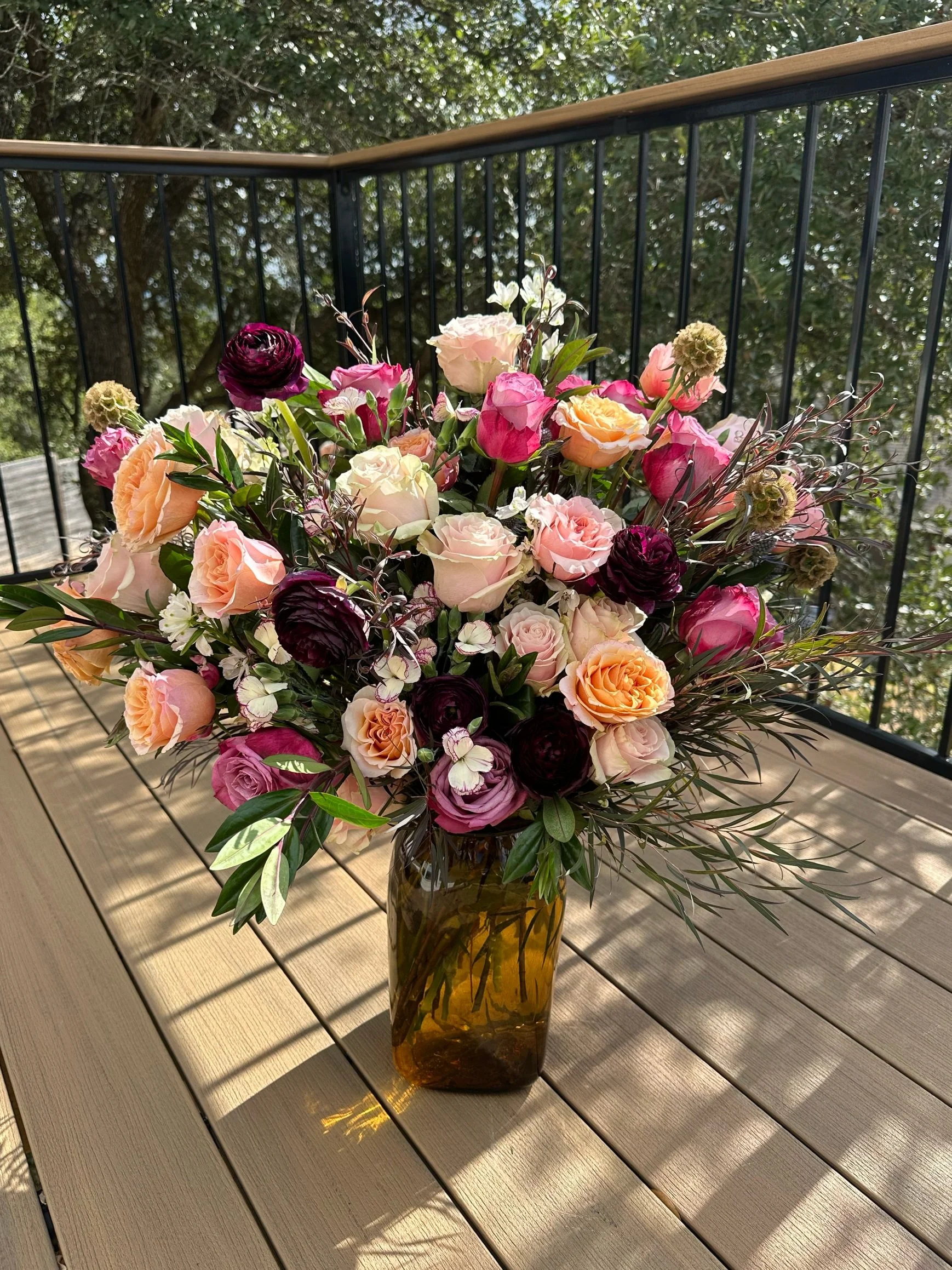 A bouquet of various pink, peach, cream, and dark purple flowers in a tall amber-colored glass vase on a wooden outdoor table, with a background of trees and a railing.