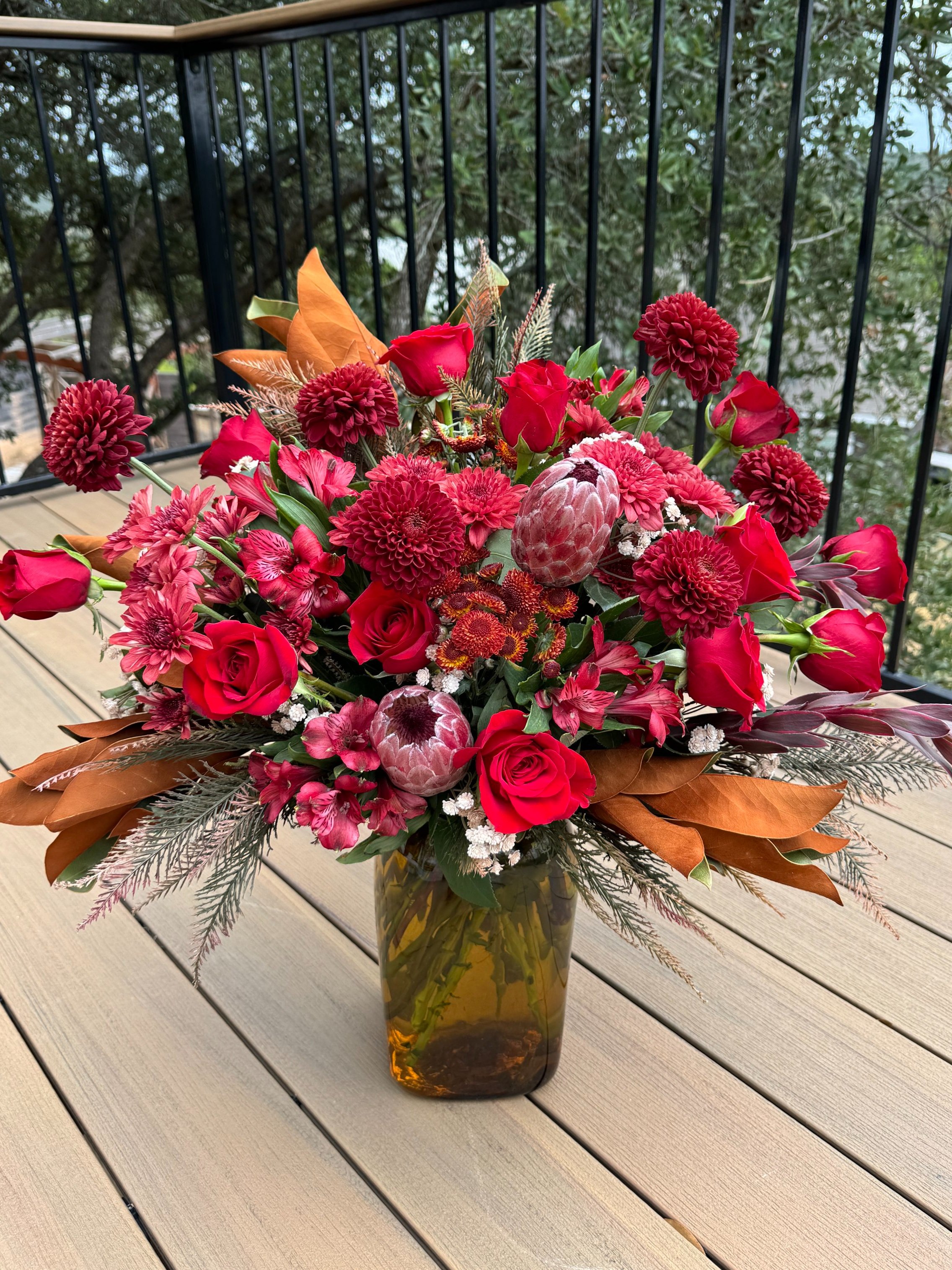 A bouquet of red flowers, including roses and dahlias, decorated with brown and red leaves, arranged in a tall amber-colored glass vase on a wooden outdoor table, with trees in the background.