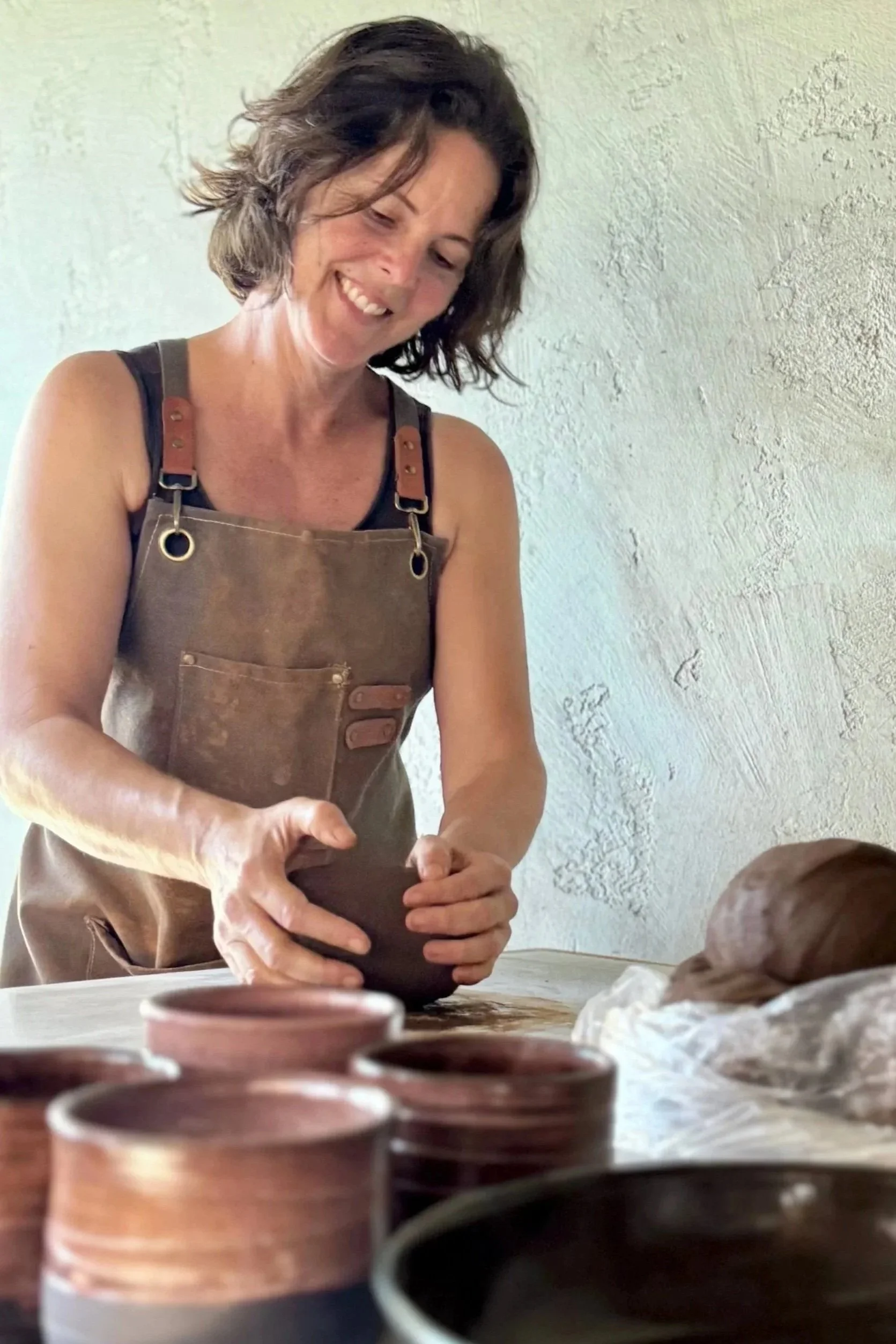 Woman with shoulder-length brown hair, smiling, wearing a brown apron over a black top, working with clay in a pottery studio.