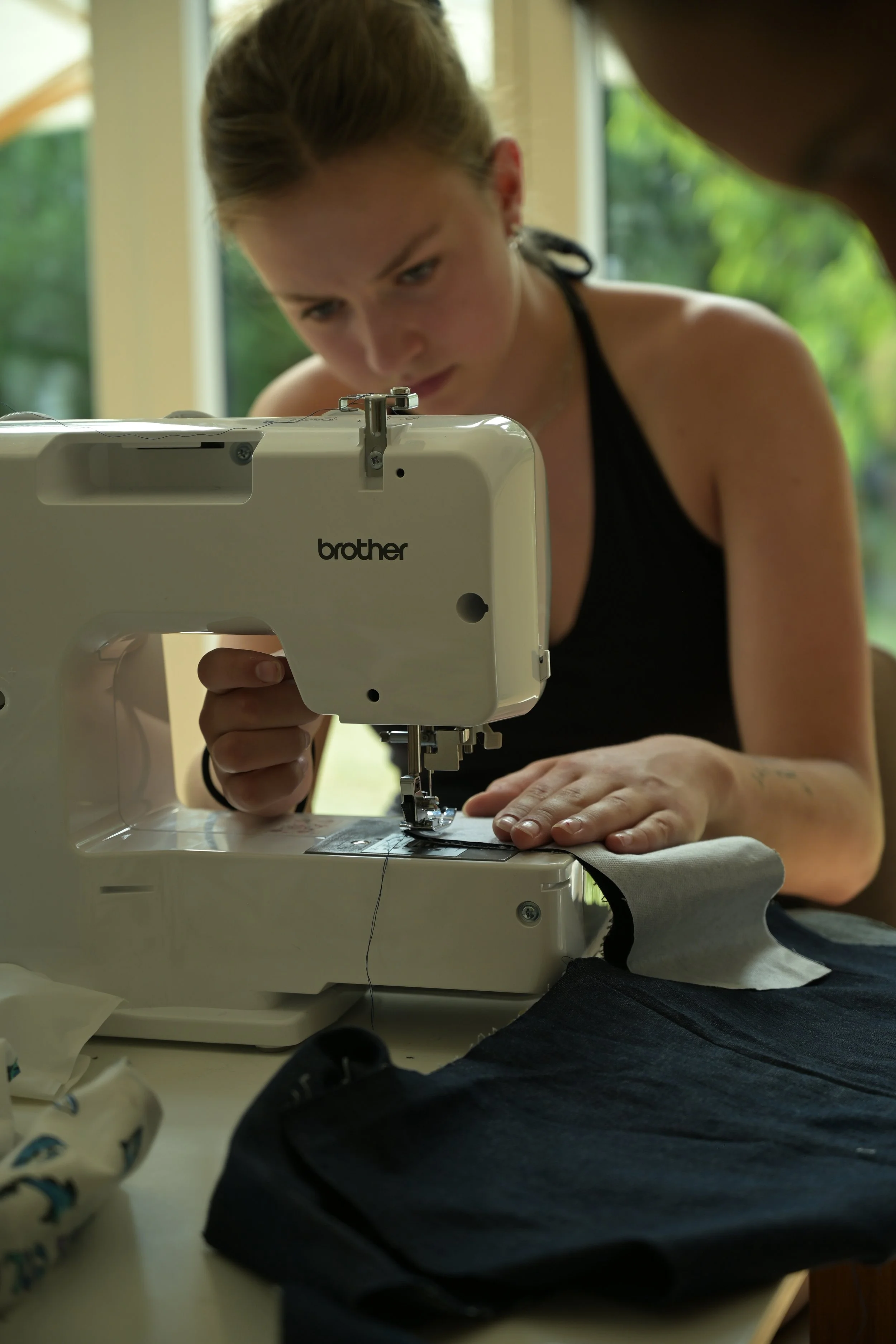 A woman sewing fabric with a Brother sewing machine, wearing a black top and sitting at a table, with a window and green scenery in the background.