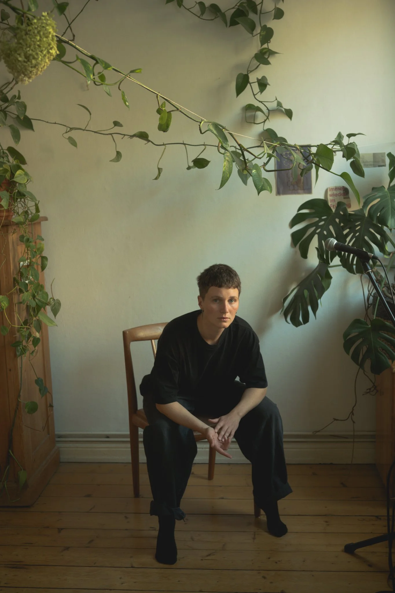A person with short hair and dark clothing sitting on a wooden chair in a room with green plants and wooden flooring.