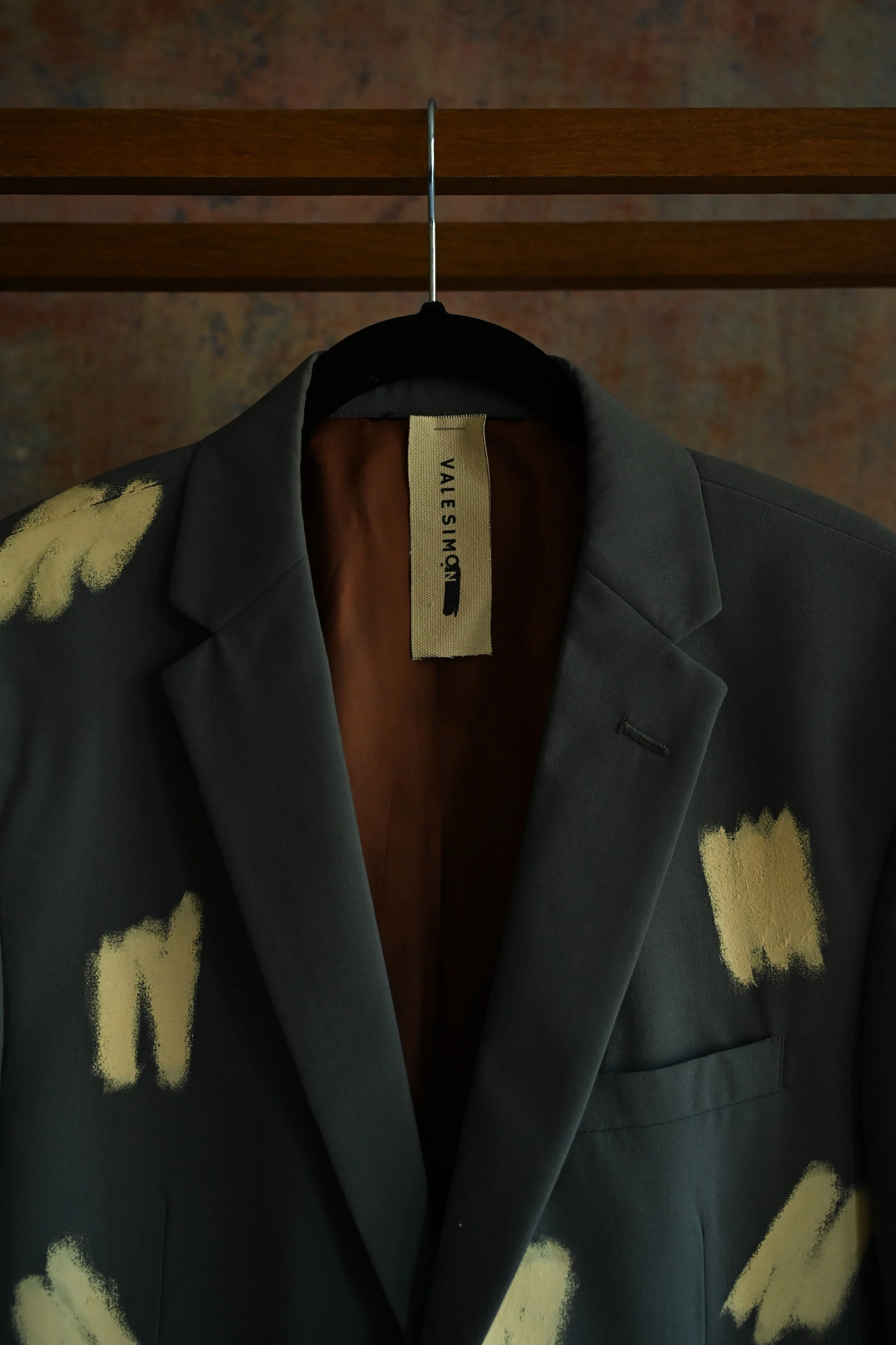 A black blazer with yellow painted or embroidered floral designs hanging on a metal hanger, with a wooden rack in the background.