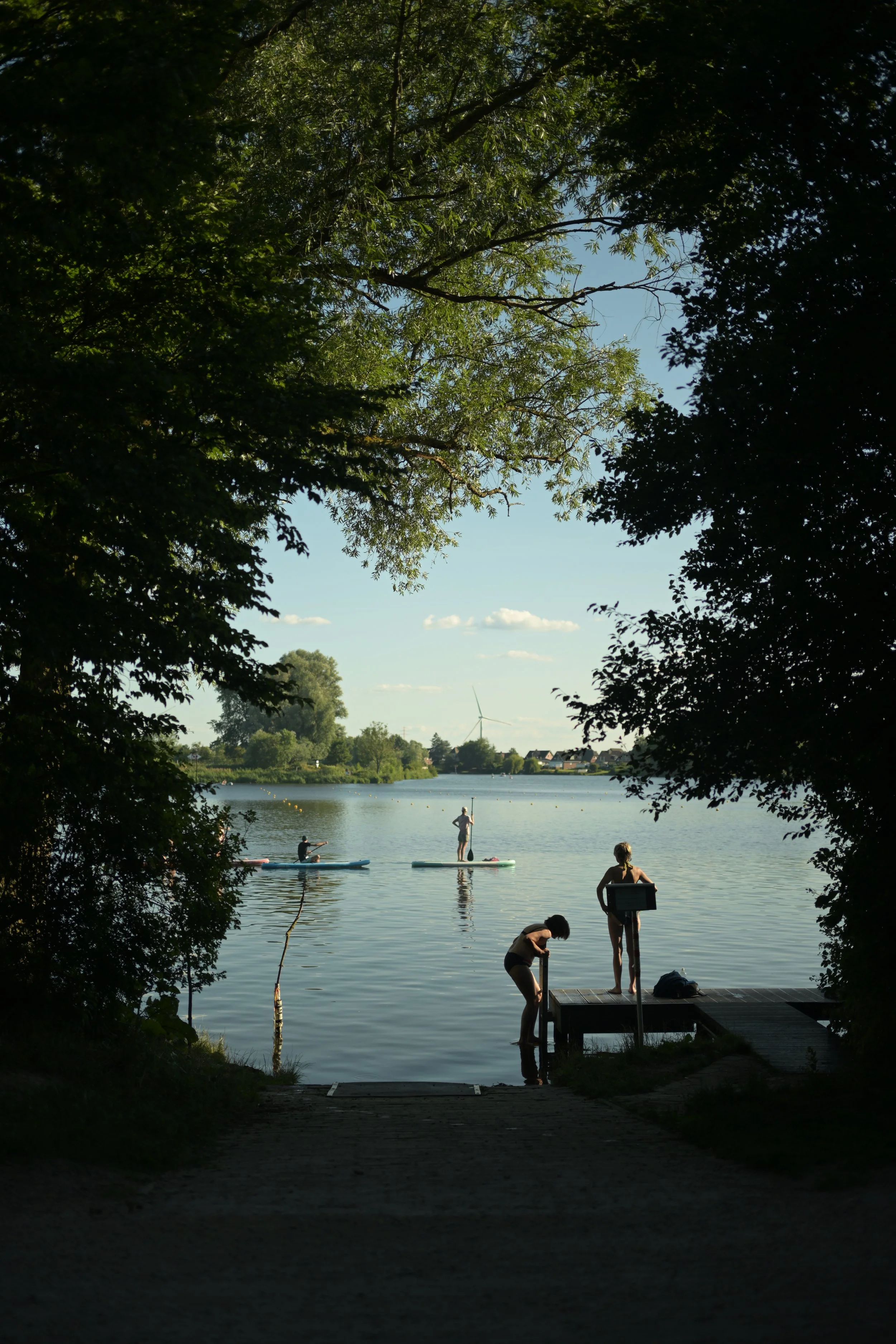 People paddleboarding, swimming, and relaxing by a lake, with trees framing the scene and a wind turbine visible in the background on a sunny day.
