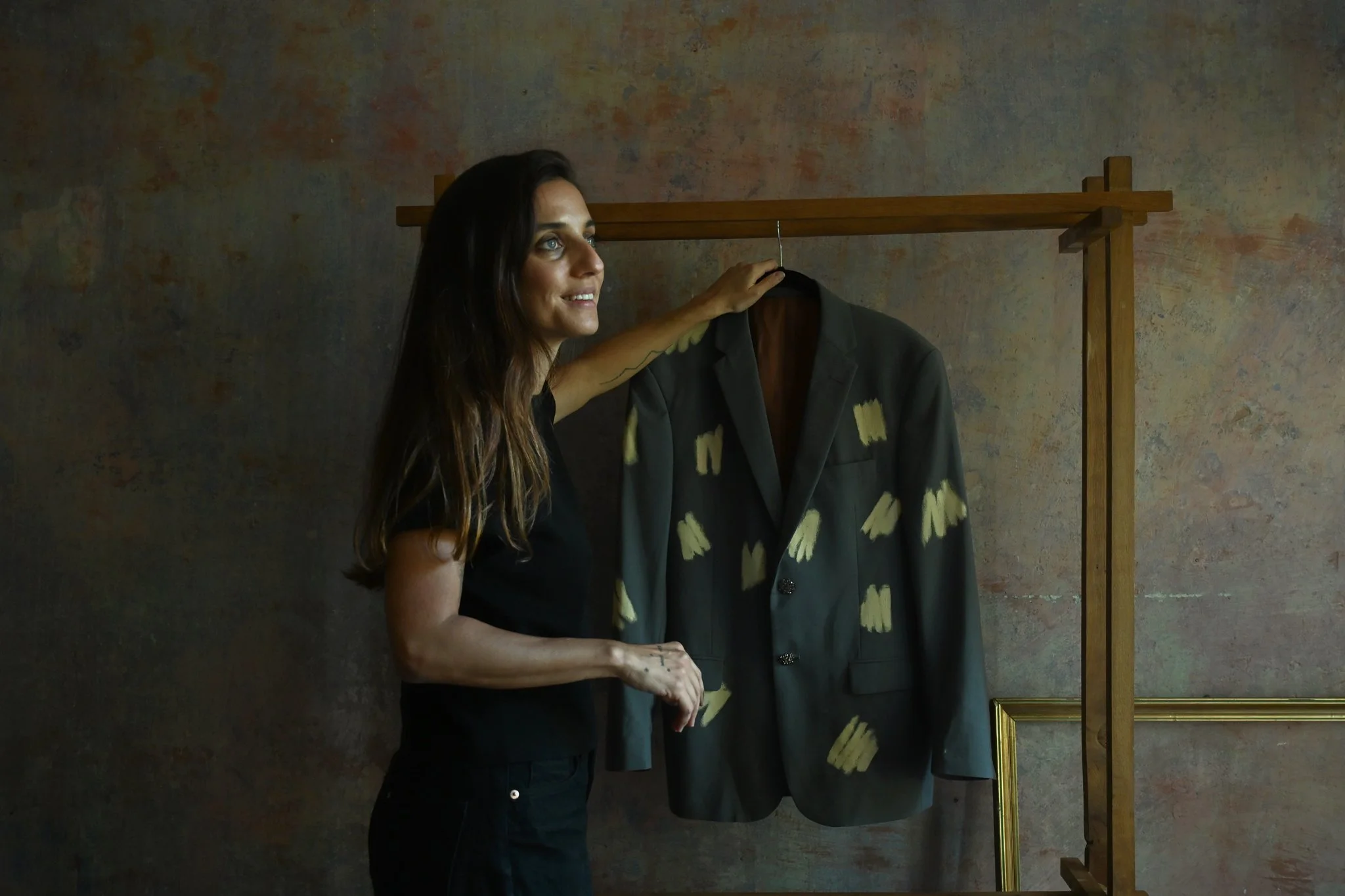 A woman with long dark hair and light eyes stands next to a clothing rack holding a shirt with yellow patterns. She is smiling and touching the shirt with her hand. The background is a textured wall with warm tones.