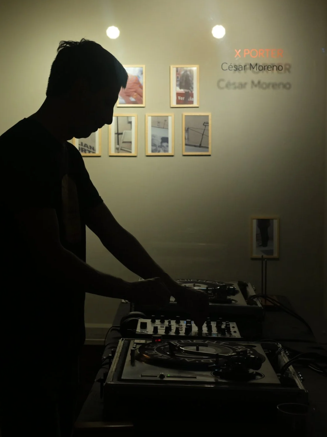 Silhouette of a DJ mixing music at a dimly lit art gallery or exhibition space. Framed photographs are displayed on the wall behind him, and a projection on the wall shows the text 'X PORTER César Moreno'.