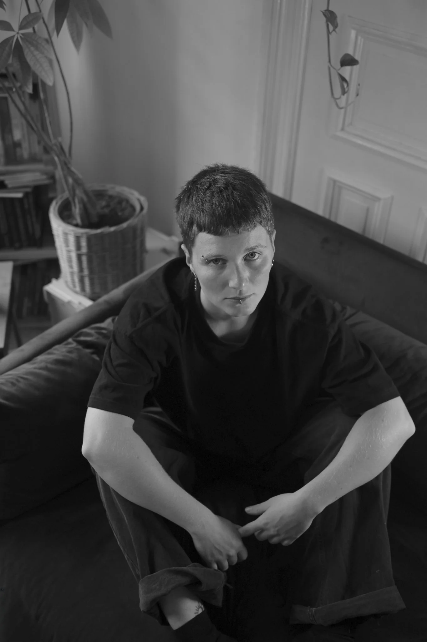 A black and white photo of a person with short hair, tattoos, and facial piercings, sitting on a bed with arms crossed over knees, looking directly at the camera in a room with bookshelves, a plant, and a cabinet.