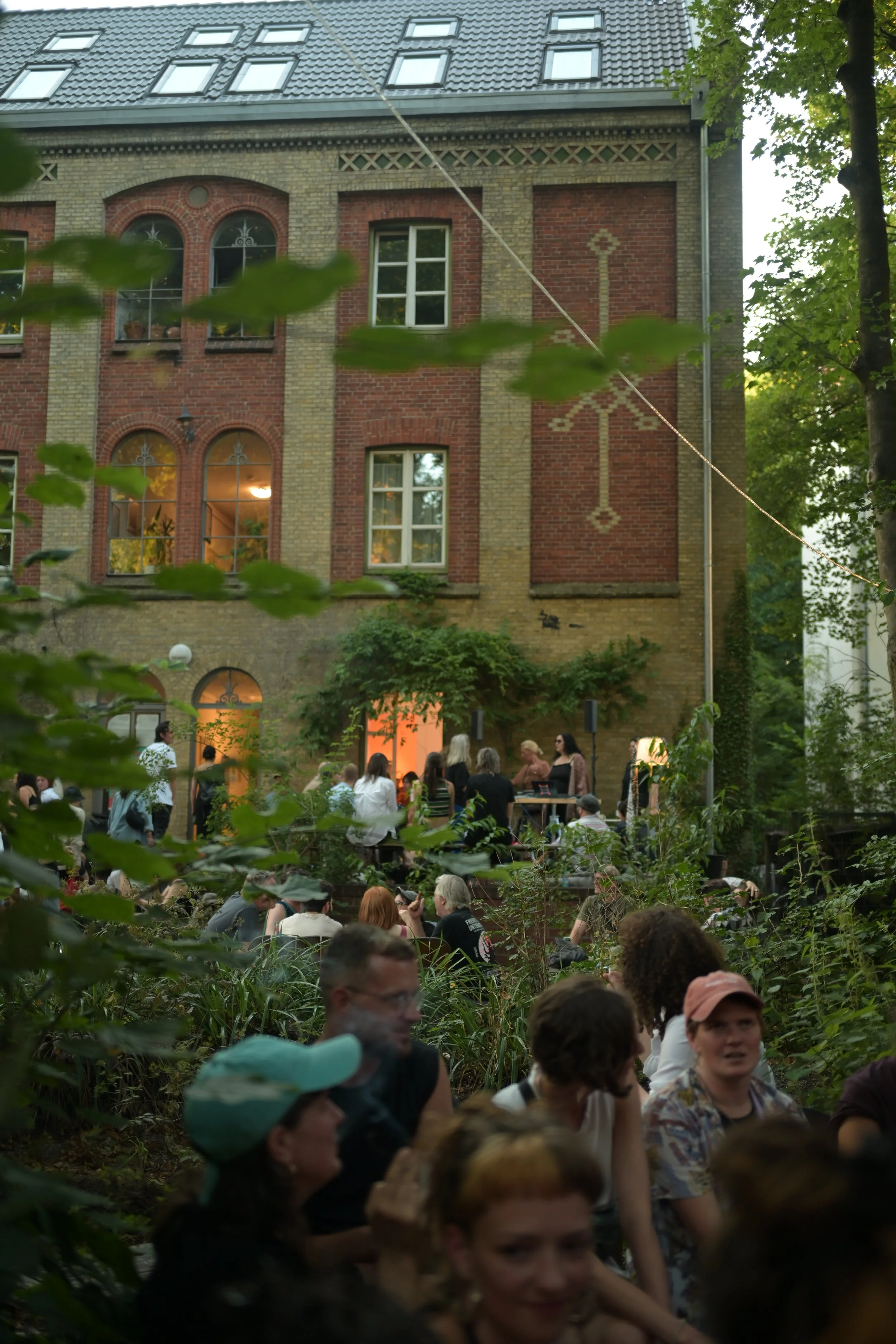A crowd gathered outdoors in front of a brick building with large windows, surrounded by green trees and foliage, during daylight.
