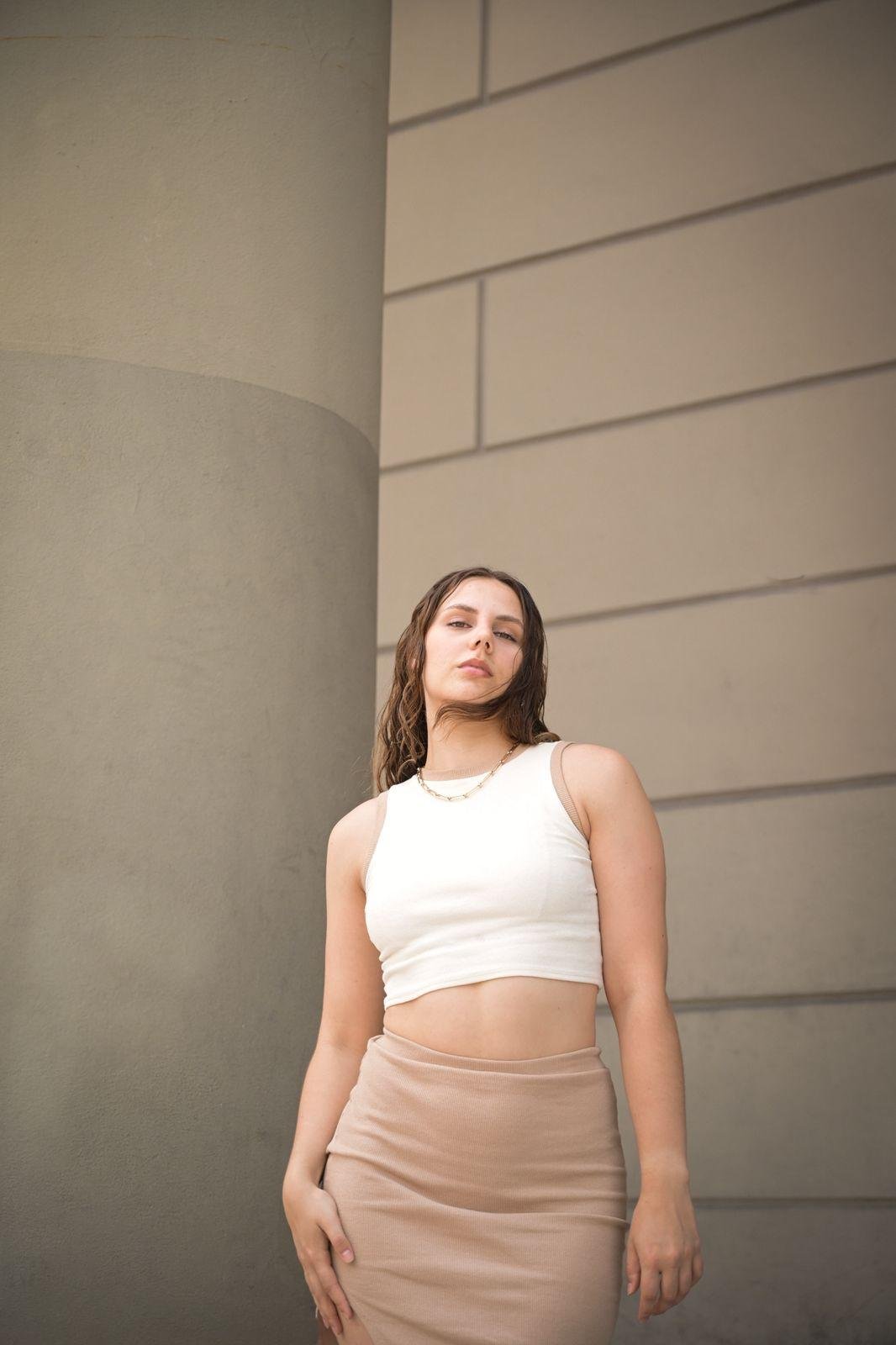 A woman with wavy brown hair poses against a beige wall and column, wearing a white cropped tank top and a beige skirt, looking confidently at the camera.