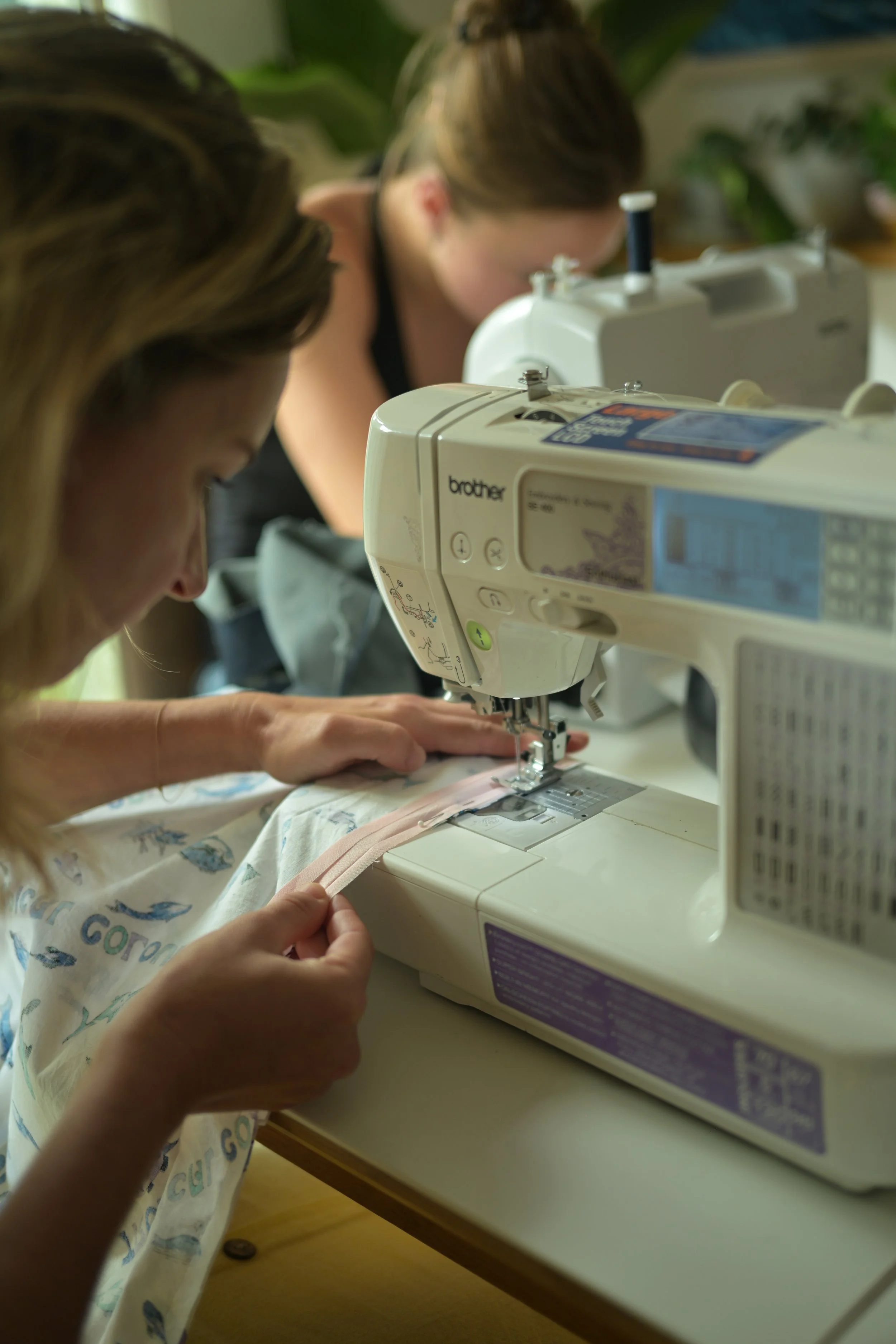 Two women are sewing fabric on sewing machines, one in the foreground wearing a colorful shirt and the other in the background wearing a dark top. They are working at a table with green plants and a shelf with books in the background.