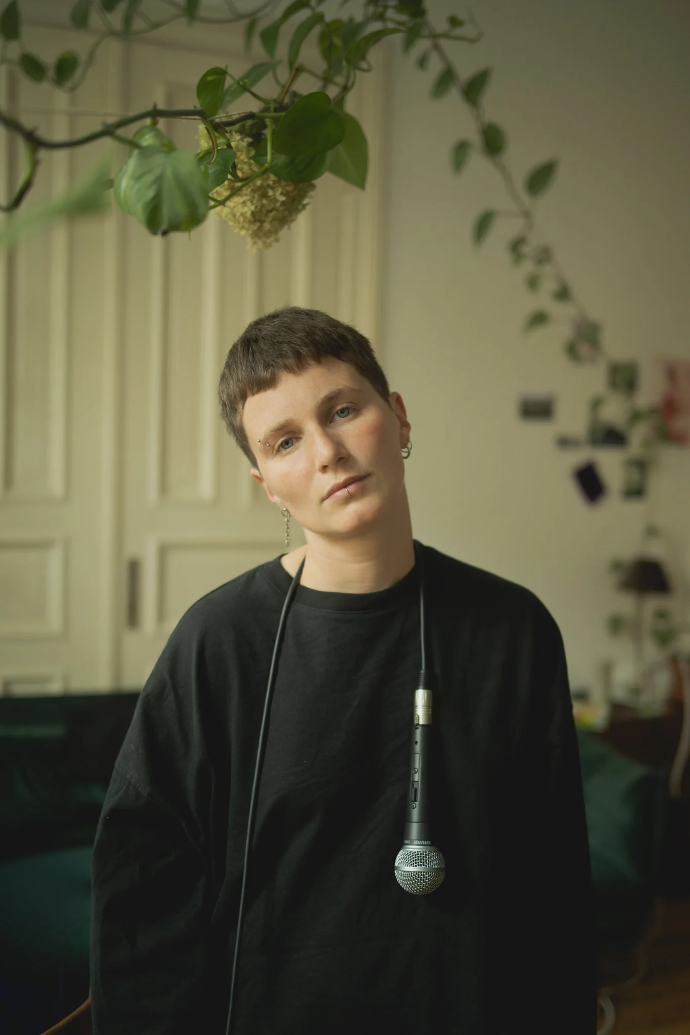 A person with short hair and earrings holding a microphone, standing in a room with photo and paper decorations, and hanging plant.