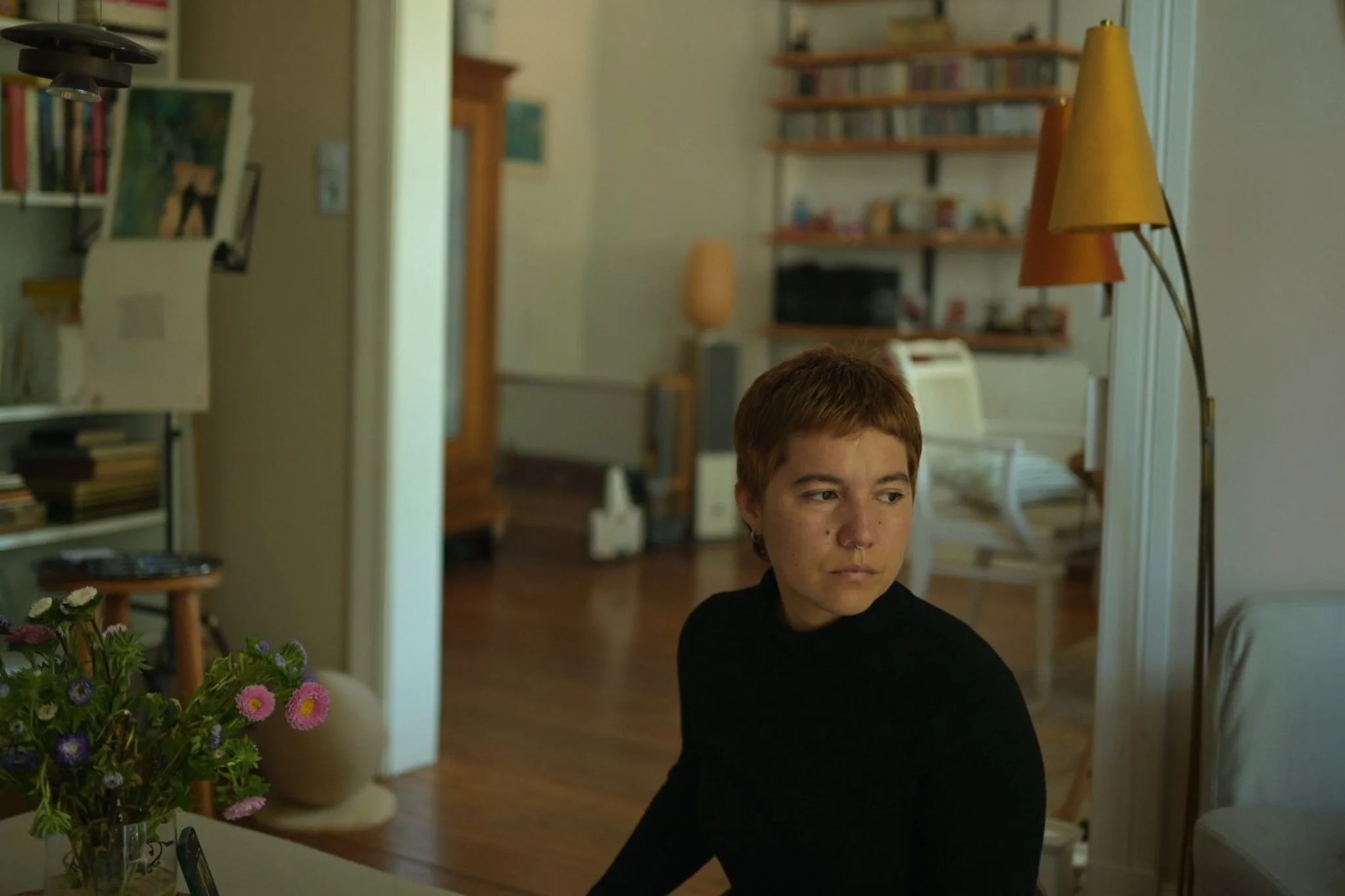 A person with short reddish hair and a septum piercing sitting in a living room, with a bookshelf, a lamp, and a microwave in the background, and a bouquet of pink, purple, and white flowers in the foreground.
