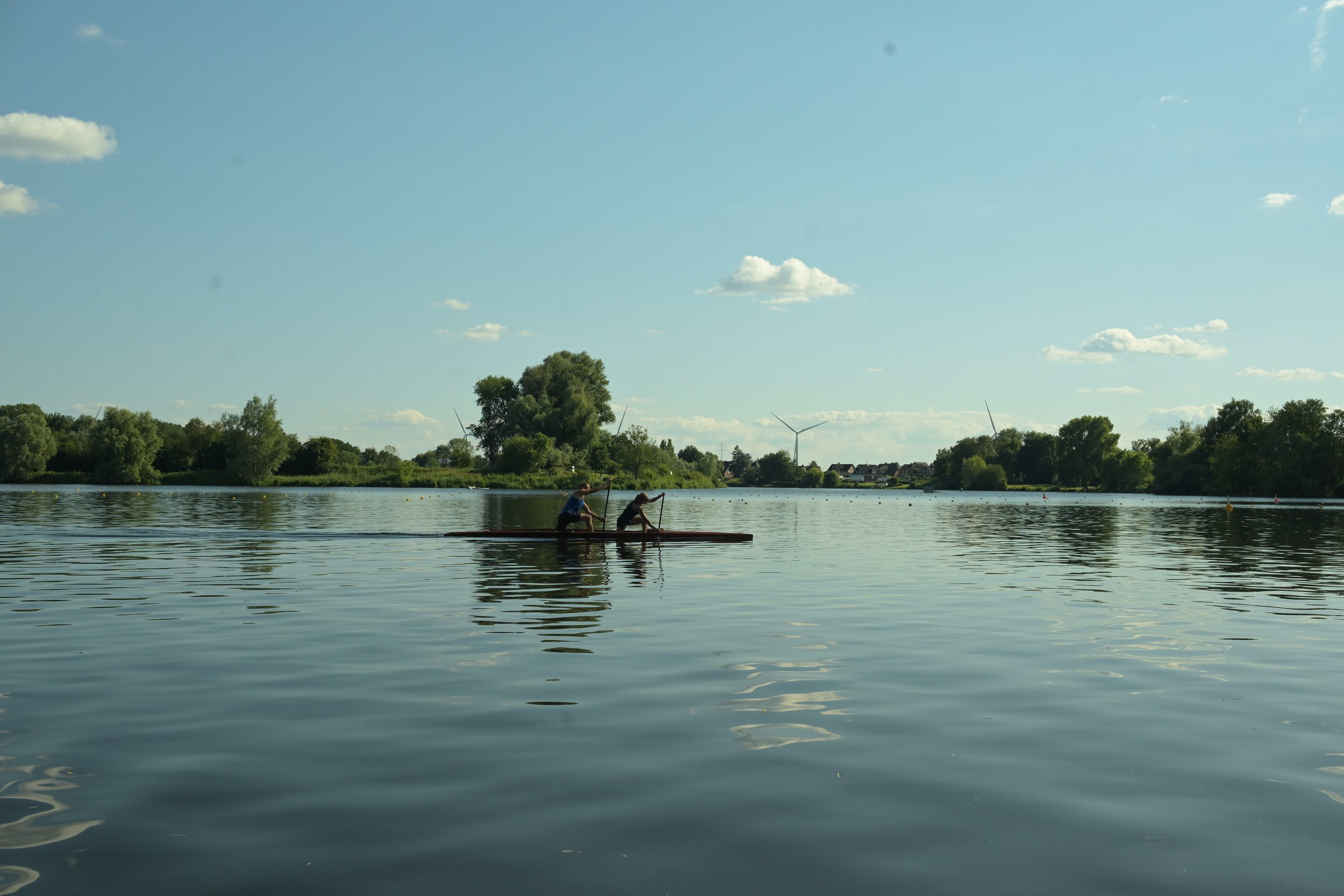 Two people on a paddleboard in a calm lake with trees and wind turbines in the background on a sunny day with blue sky and scattered clouds.
