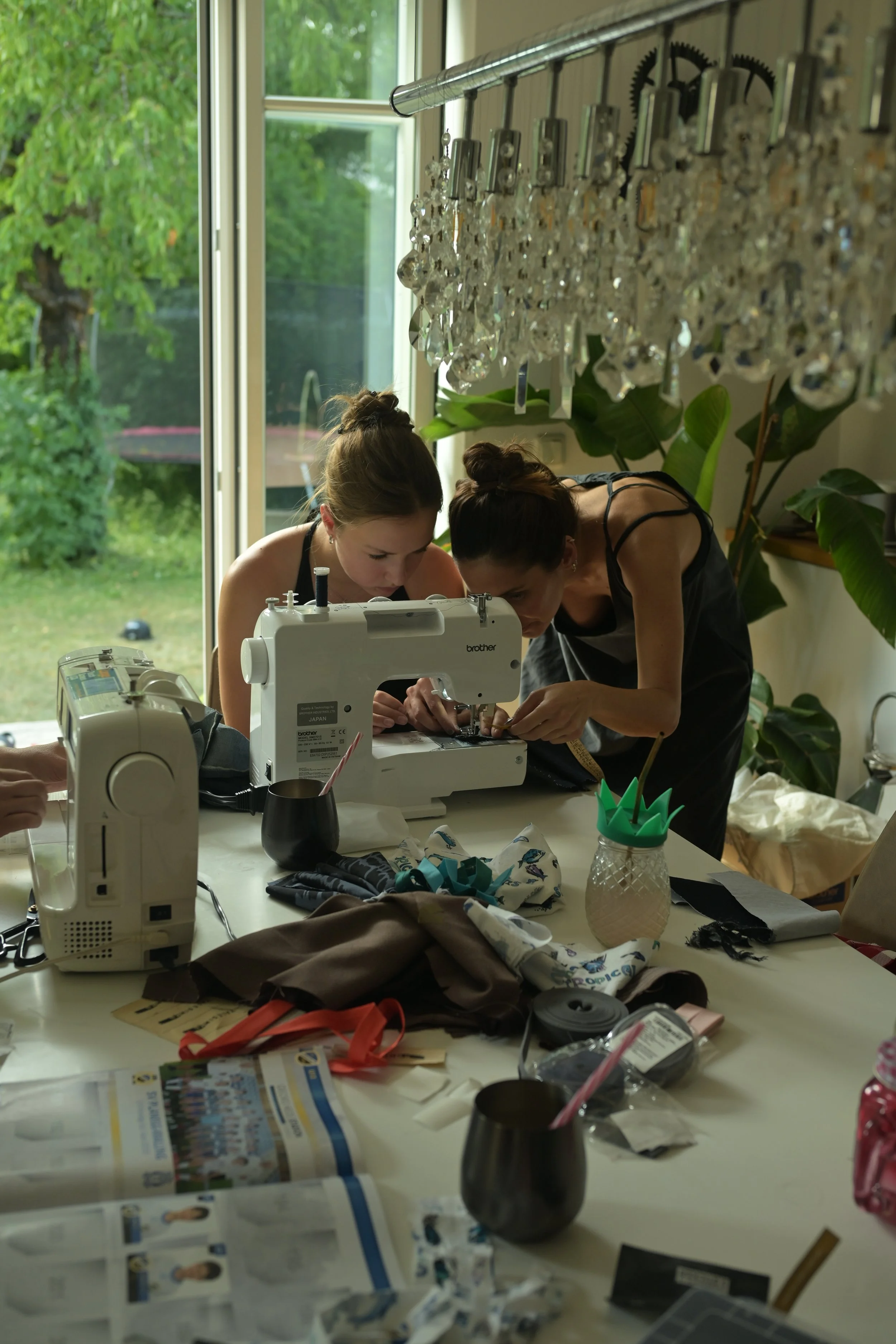 Two women working together on a sewing project at a cluttered table with sewing machines, fabric, and tools, in a room with large windows and green plants.