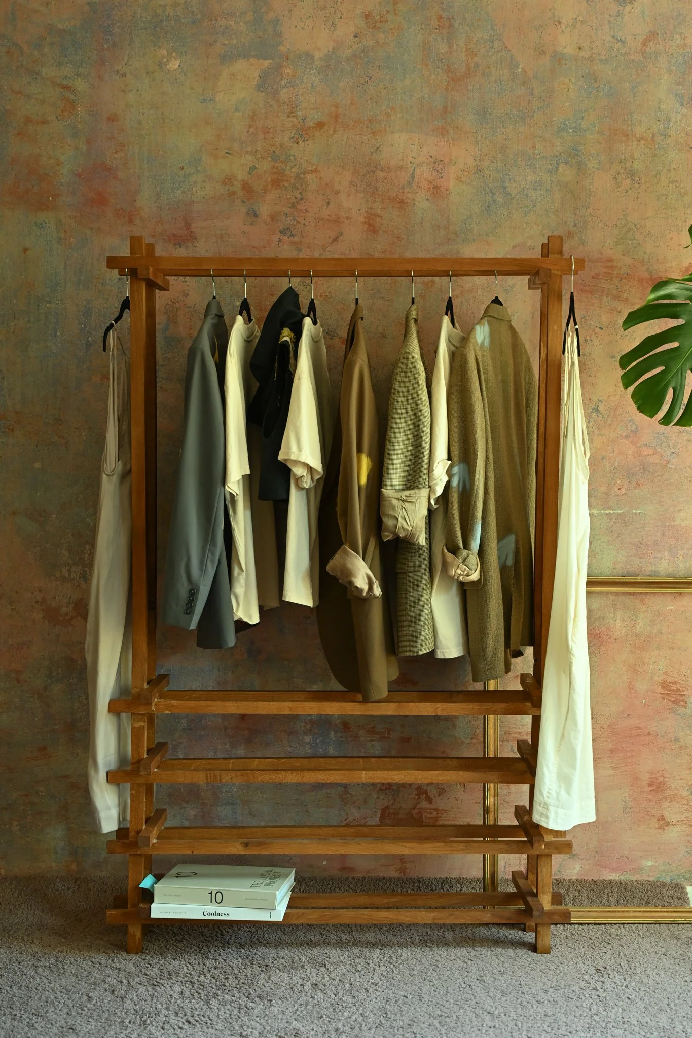 A wooden clothing rack with various shirts and jackets hanging on it, positioned against a textured wall with a small plant on the right. There are three books on the bottom shelf, one of which is titled 'Coolness.'