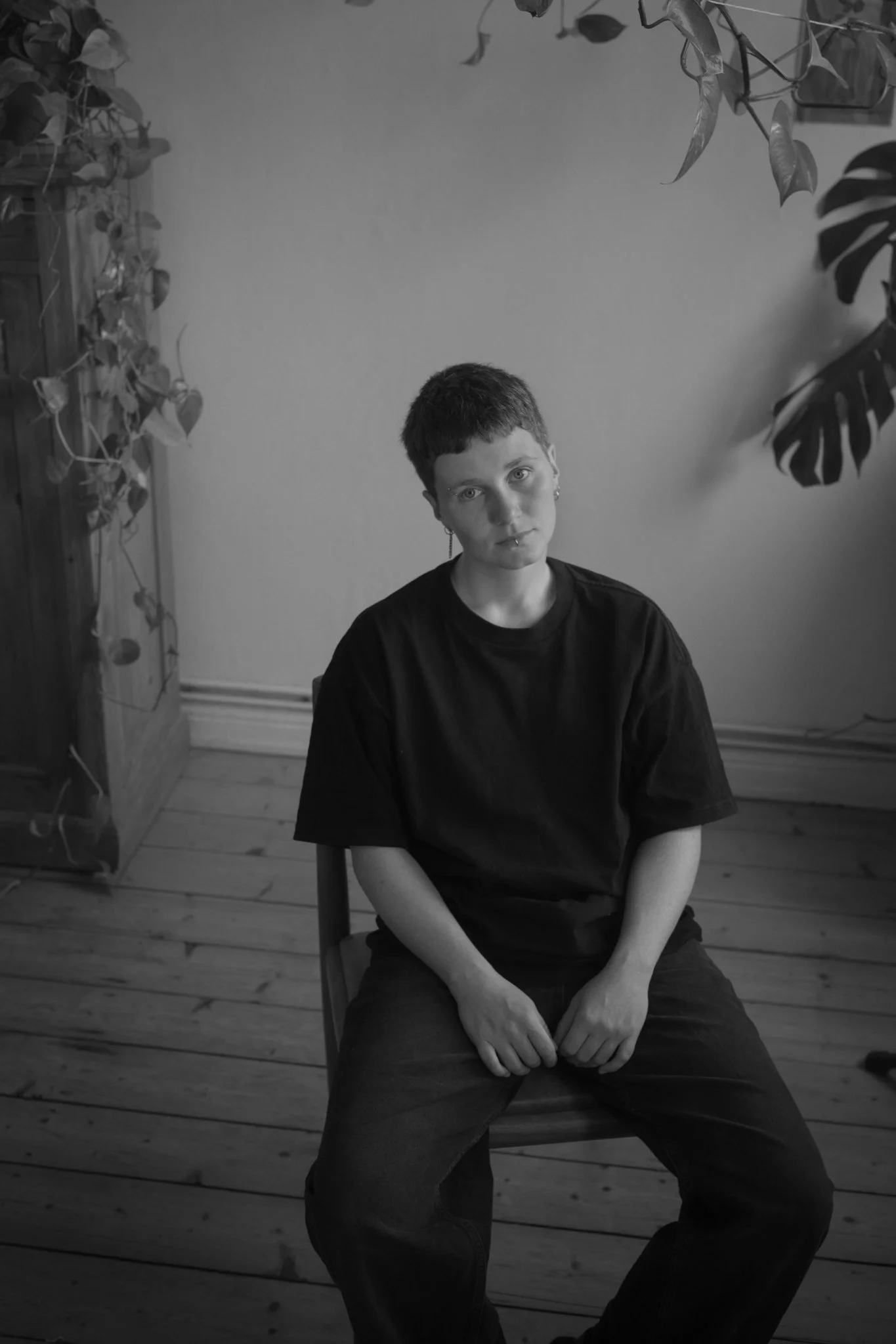 A young person with short hair and earrings sitting on a wooden chair in a room with plants and a wooden cabinet. Black and white photograph.