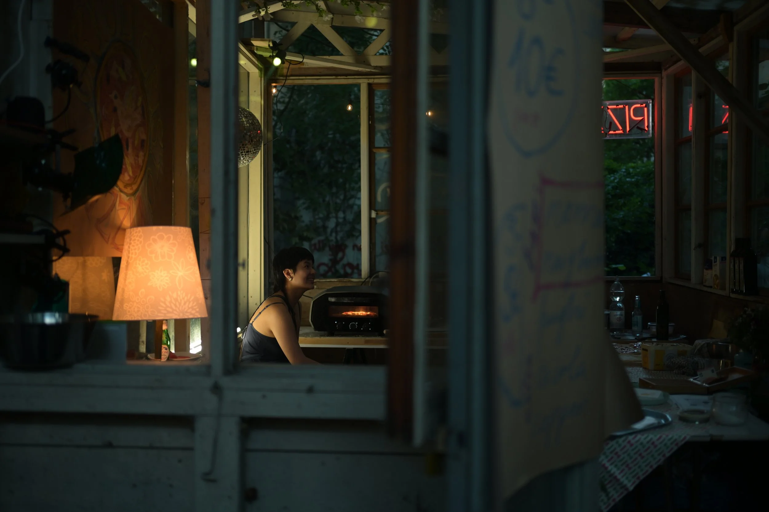 A woman sitting inside a cozy, dimly lit room with wooden walls, smiling and looking to her left. The room has warm lighting from a lamp with a floral design, a disco ball hanging from the ceiling, and various decorations including a neon sign in the window. The scene is viewed through a window with metal bars.