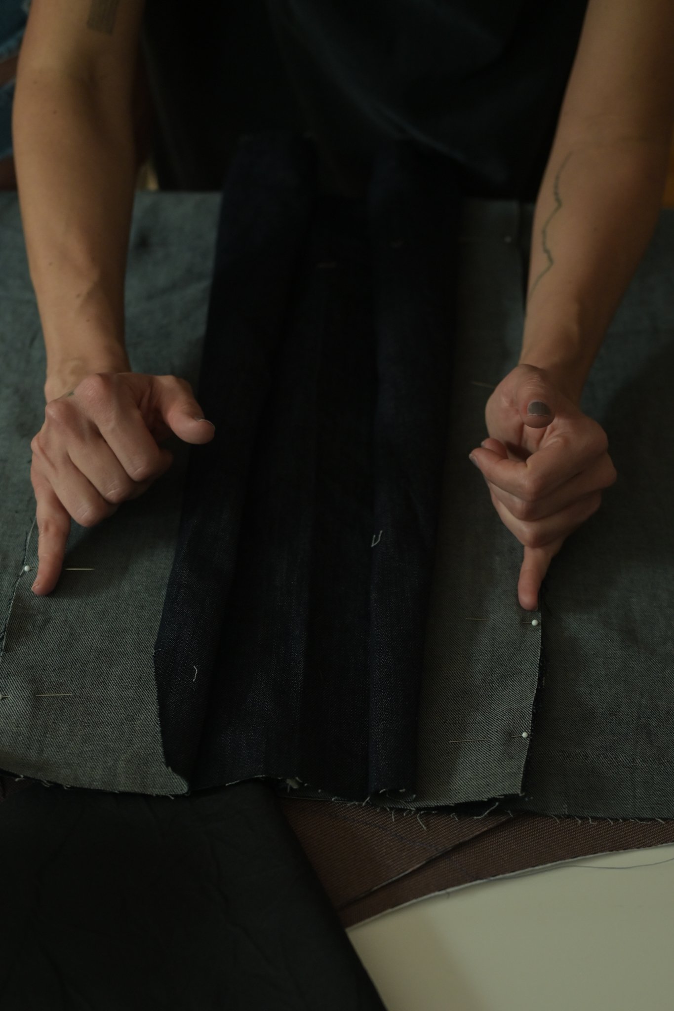 A person with light skin is sitting with their arms on their legs, wearing a black sleeveless top and dark pants. They have a tattoo on their right forearm and painted nails. The person is pointing down with both index fingers.