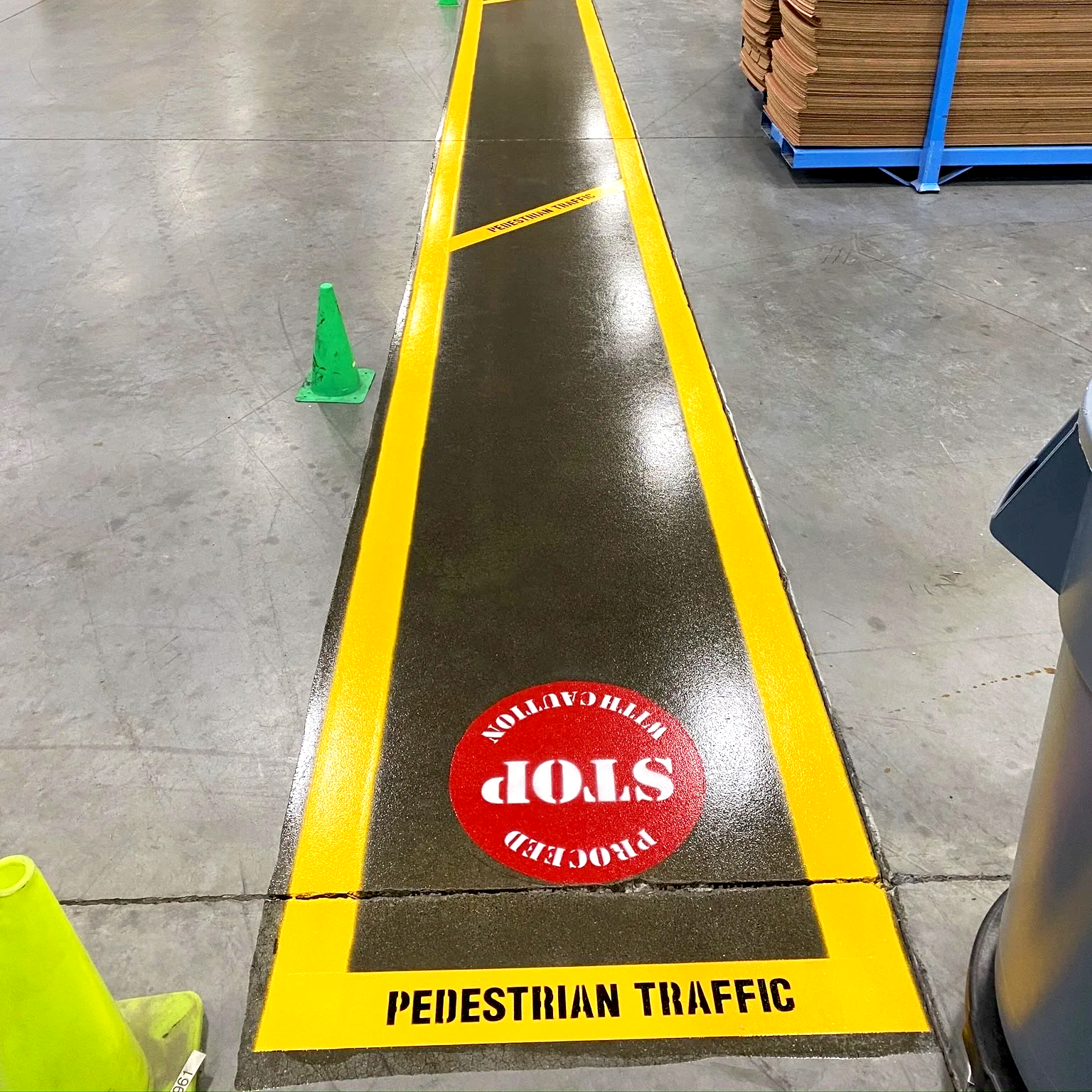 A black and yellow pedestrian traffic line on the floor with a red stop sign in the middle that reads "STOP" and various caution and warning words. The line is in an industrial or warehouse setting with concrete floors and stacked cardboard boxes in the background.