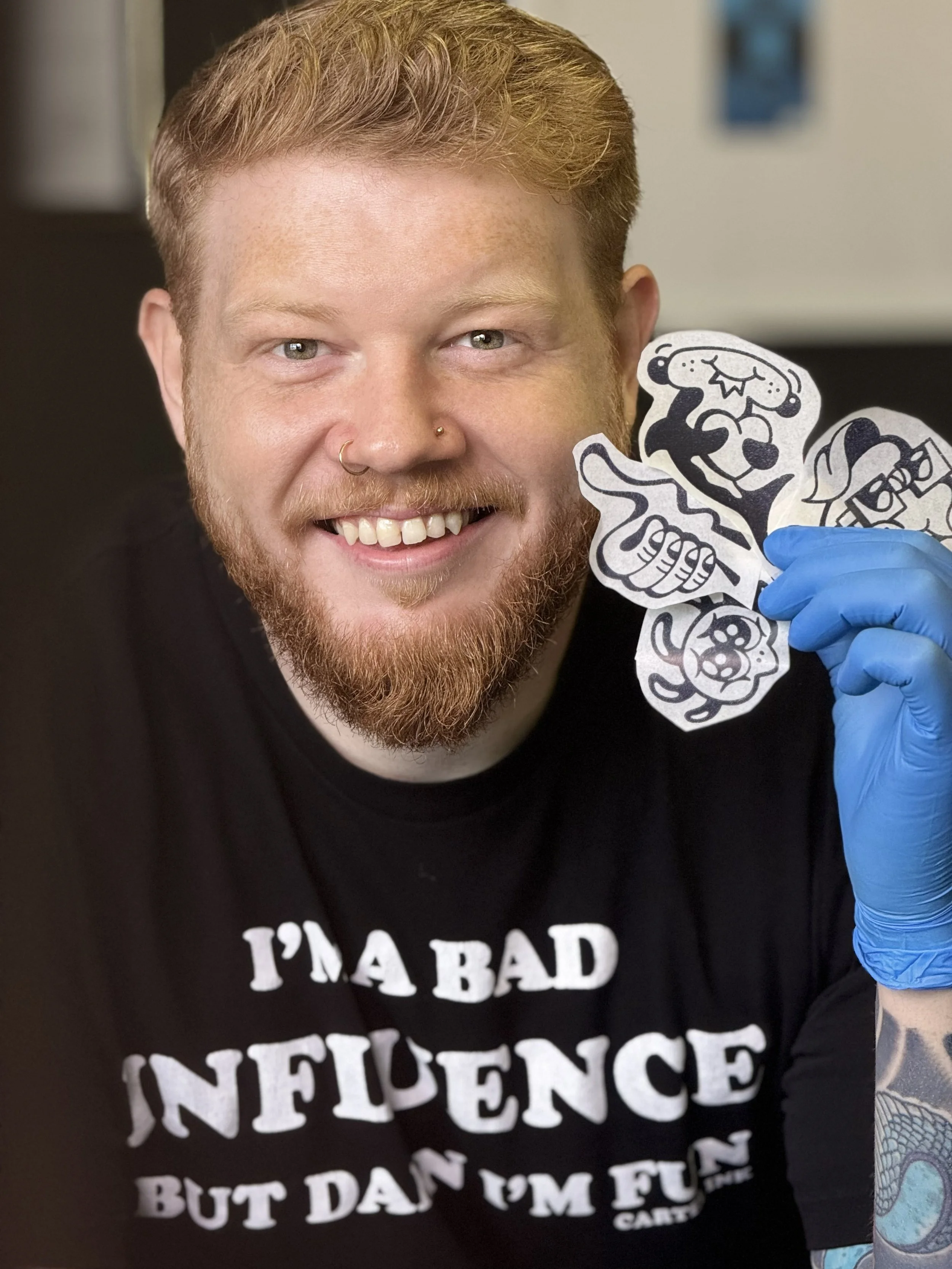 A man with a beard and mustache, wearing a black shirt with white text, and a nose ring, holding paper cutouts of cartoon creatures. The background is indoor with blurred posters or pictures.