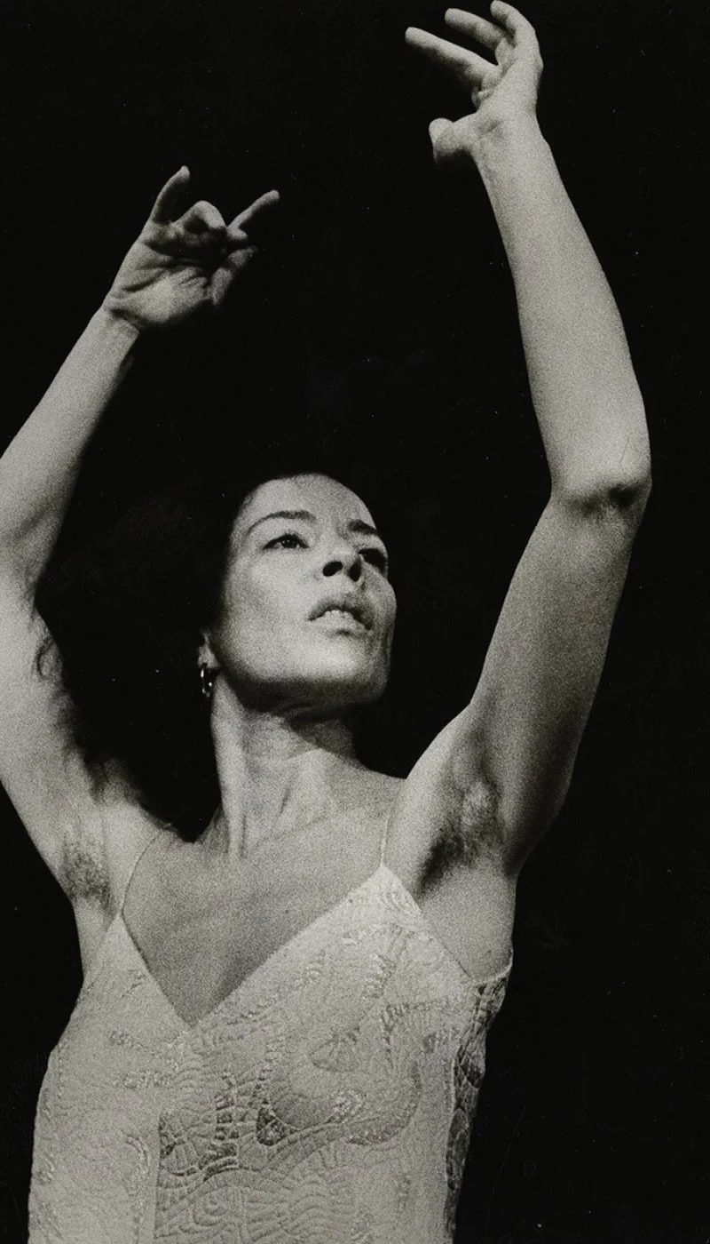 Black and white photo of dancer Deborah Hay, wearing a sleeveless dress, with her arms raised above her head and hands positioned elegantly, looking upward.