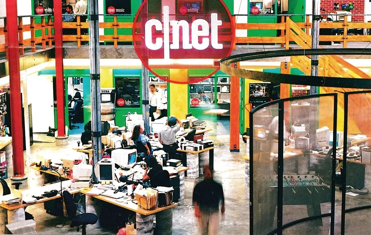 CNET office workspace with desks, computers, and people working, featuring a large CNET sign hanging overhead.