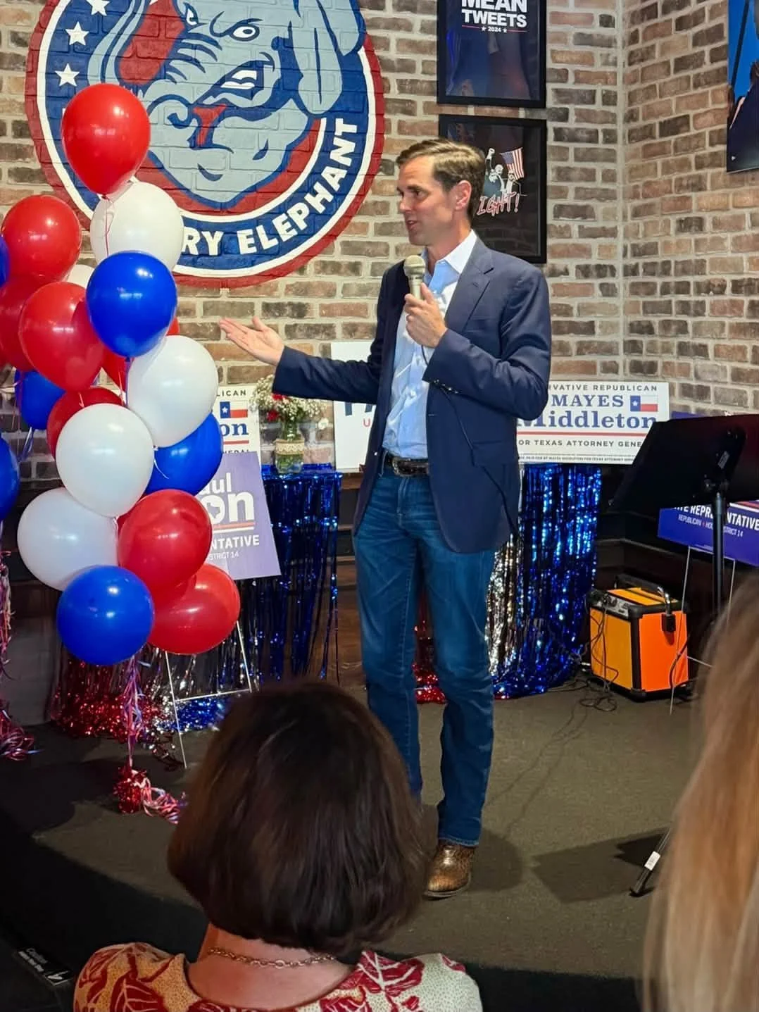 Man in a blue blazer speaking into a microphone at a political event decorated with red, white, and blue balloons, patriotic banners, and political signs, in front of a brick wall with political posters.