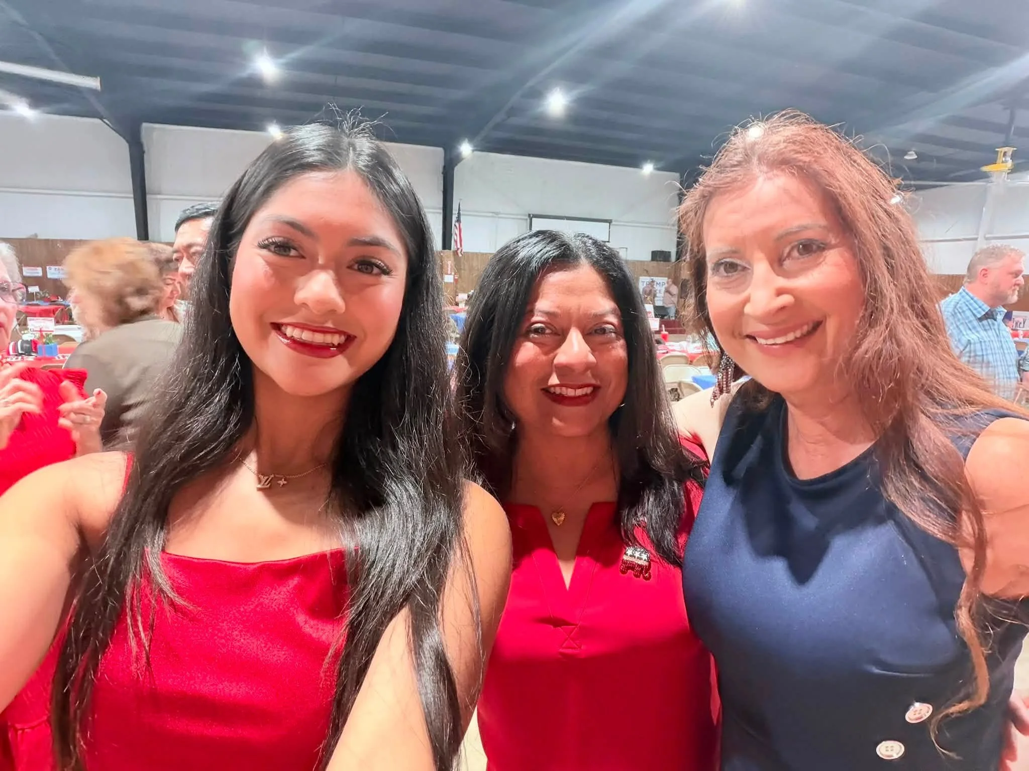 Three women smiling and taking a selfie together at an indoor event with tables, chairs, and other attendees in the background.