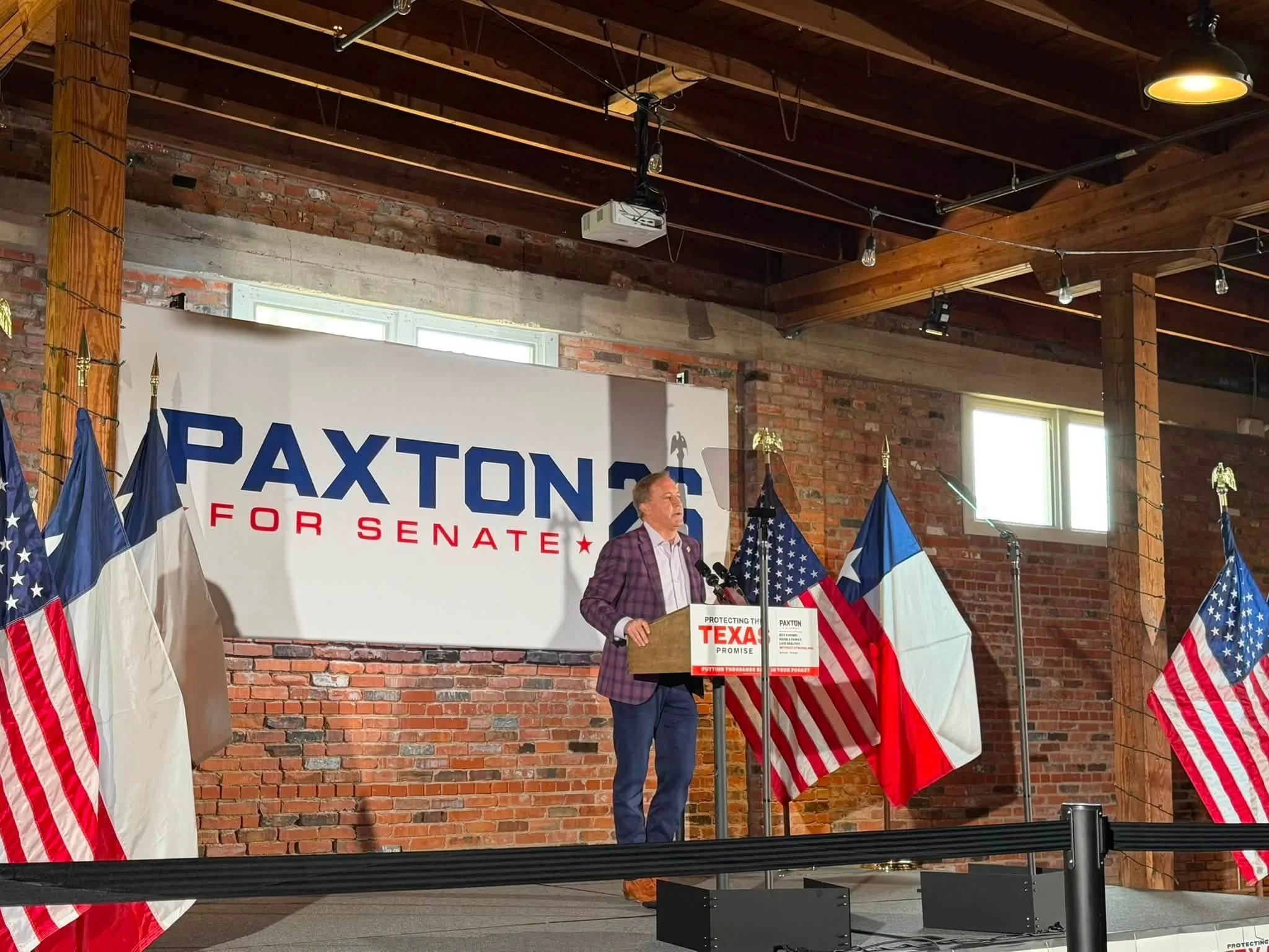 A man in a plaid blazer speaking at a podium with a sign that reads "Protecting the Texas Promise" during a political event with American flags surrounding him.
