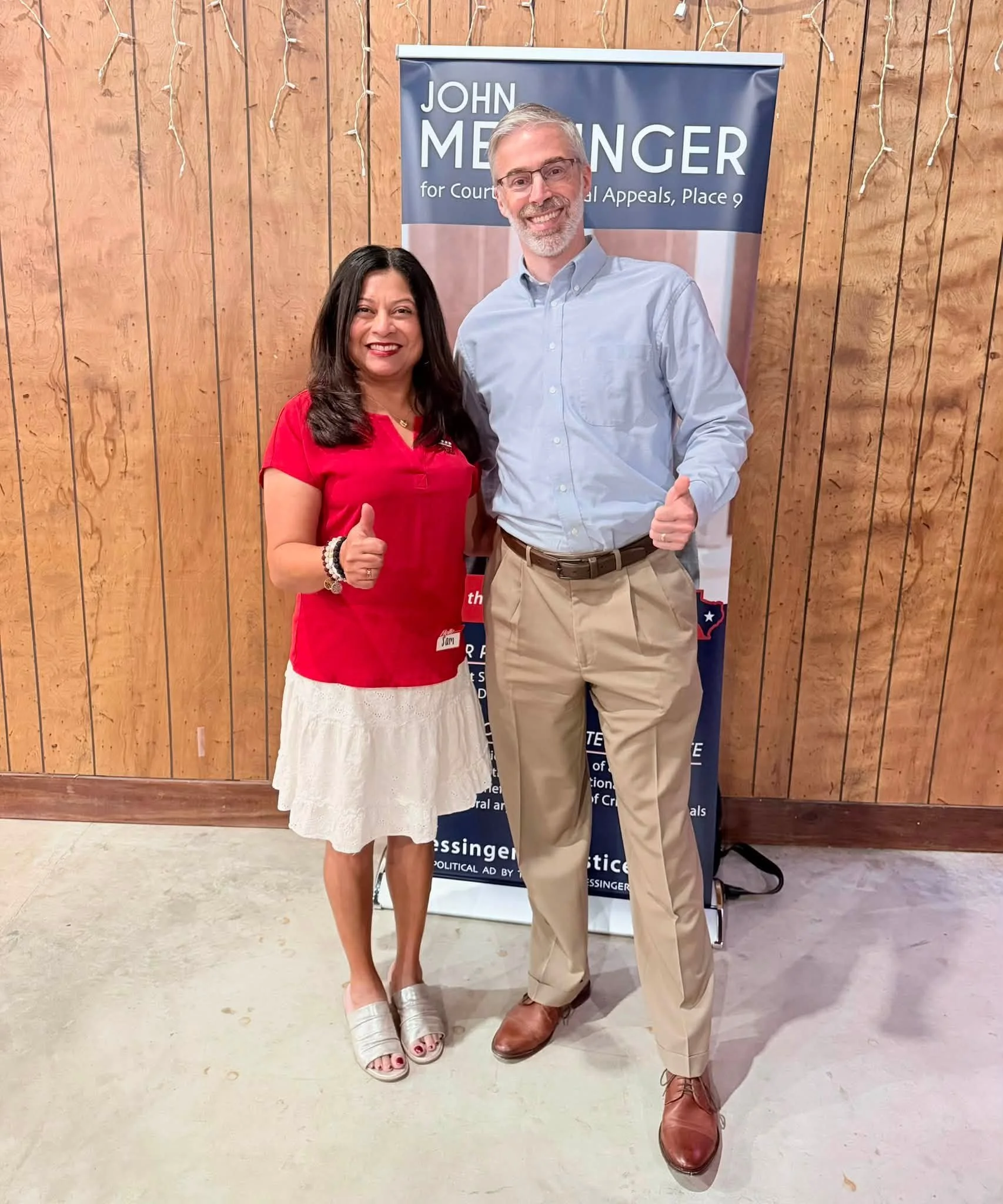 Two people smiling and giving thumbs up in front of a sign that reads "John Messenger for Court of Criminal Appeals, Place 9." The woman on the left is wearing a red shirt and a white skirt, and the man on the right is dressed in a light blue dress s
