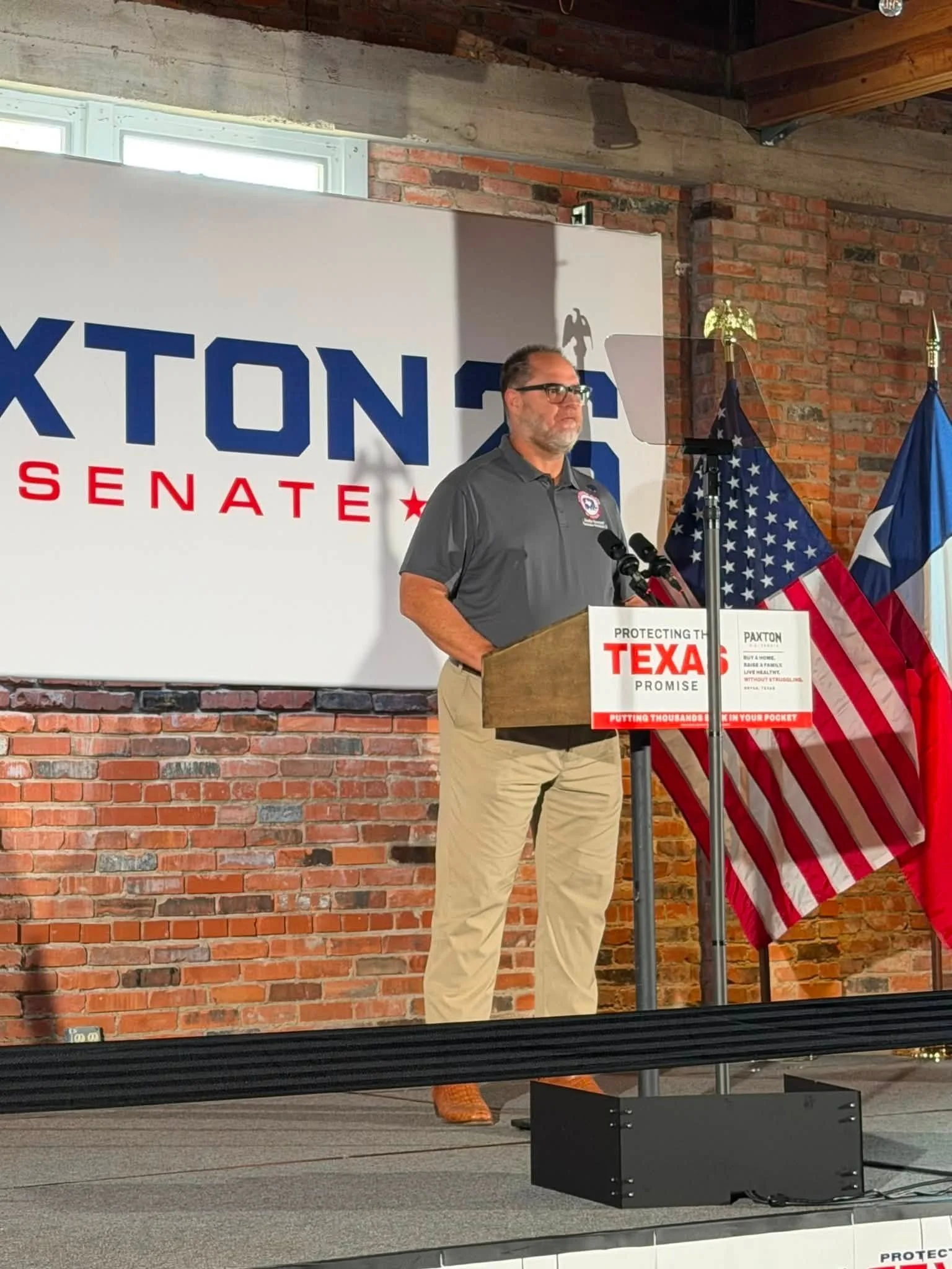 A man at a podium speaking, with a sign behind him that reads 'TEXAS' and 'SENATE' and an American flag and the Texas flag in the background. The setting appears to be a brick-walled room with windows.