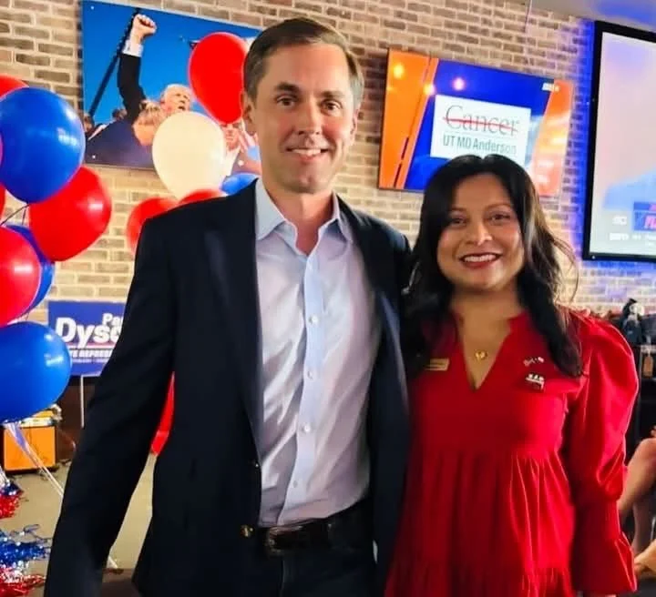 A man in a dark suit and a woman in a red dress standing together in front of a brick wall and a display with balloons and banners at a celebration or event.