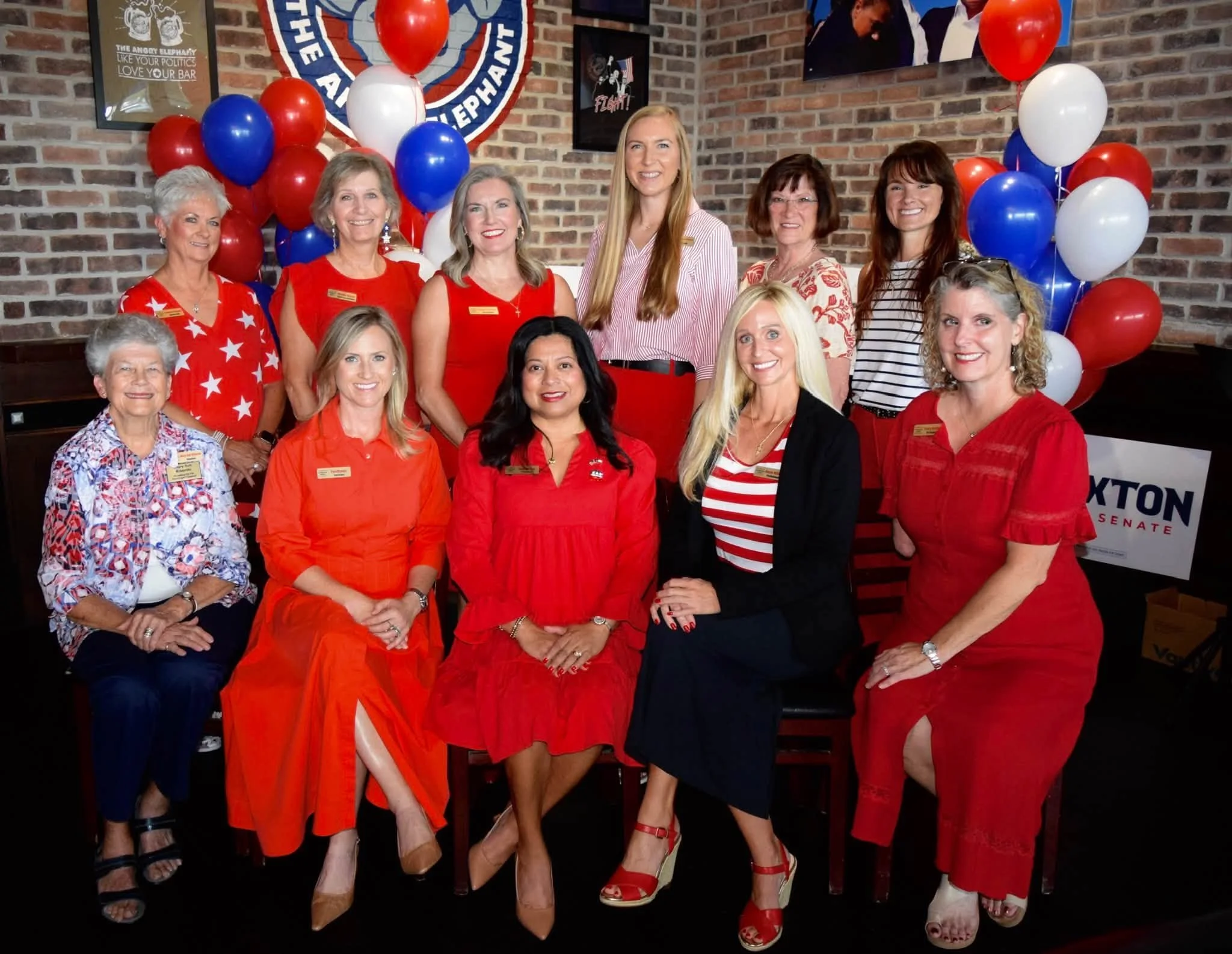 Group of women dressed in red, white, and patriotic clothing, standing together in front of a brick wall decorated with balloons and a large banner supporting a political campaign.