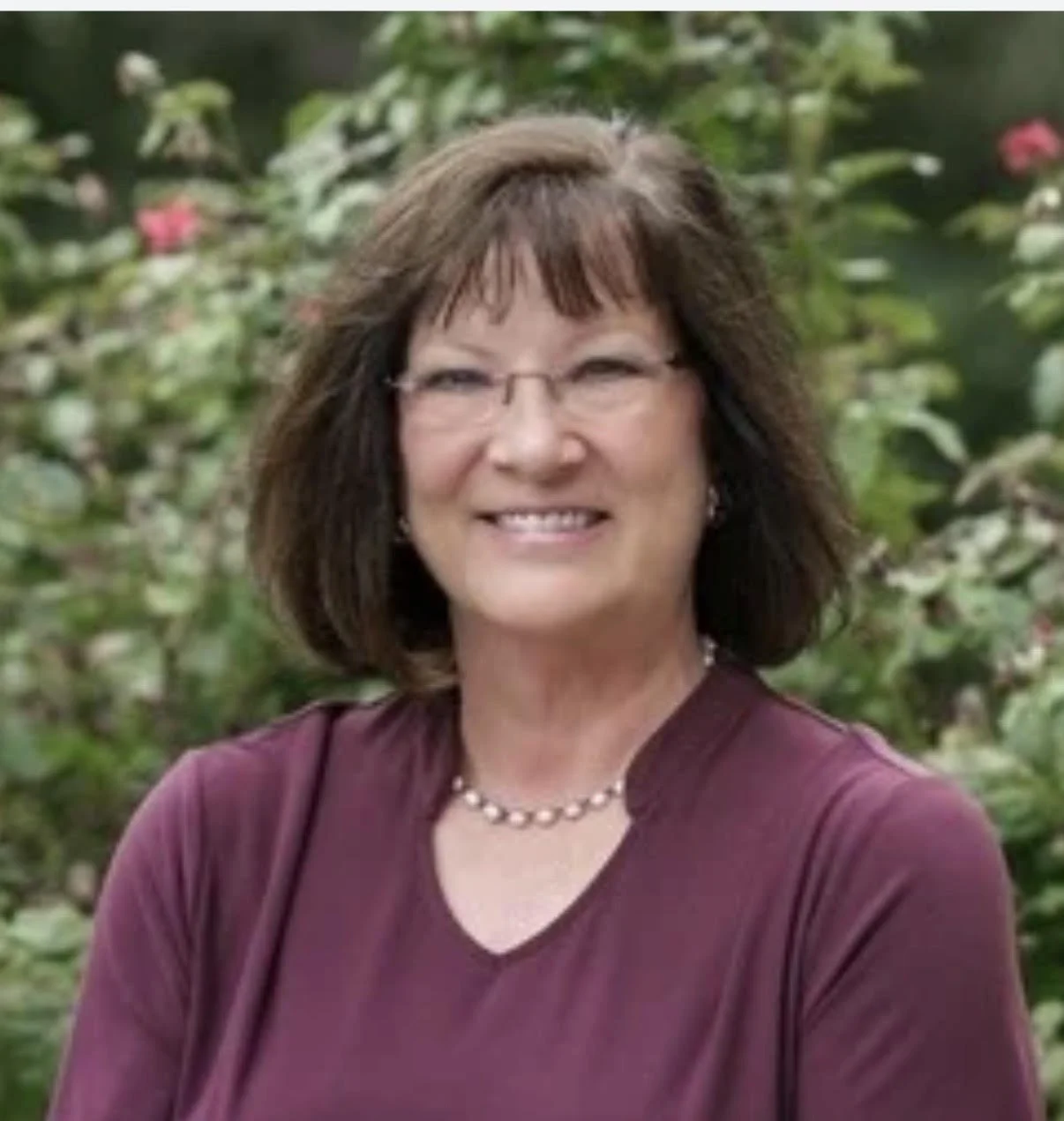 A smiling middle-aged woman with shoulder-length brown hair and glasses, wearing a maroon top and a pearl necklace, standing outdoors with greenery and flowers in the background.