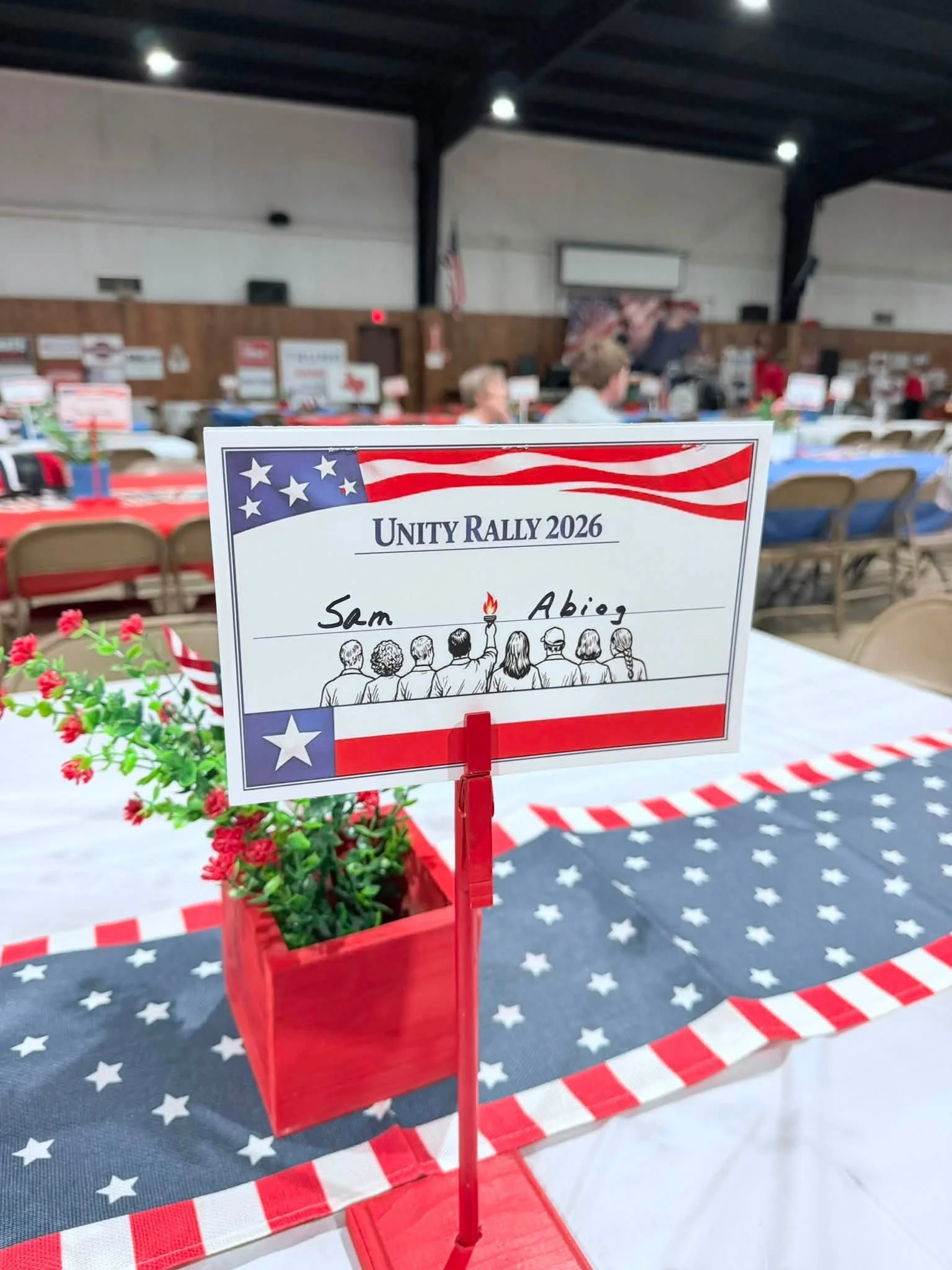 Table decorated with patriotic American flag-themed items and a sign for a Unity Rally 2026, with a red flowerpot and a backdrop of tables and chairs in a gymnasium decorated with red, white, and blue.