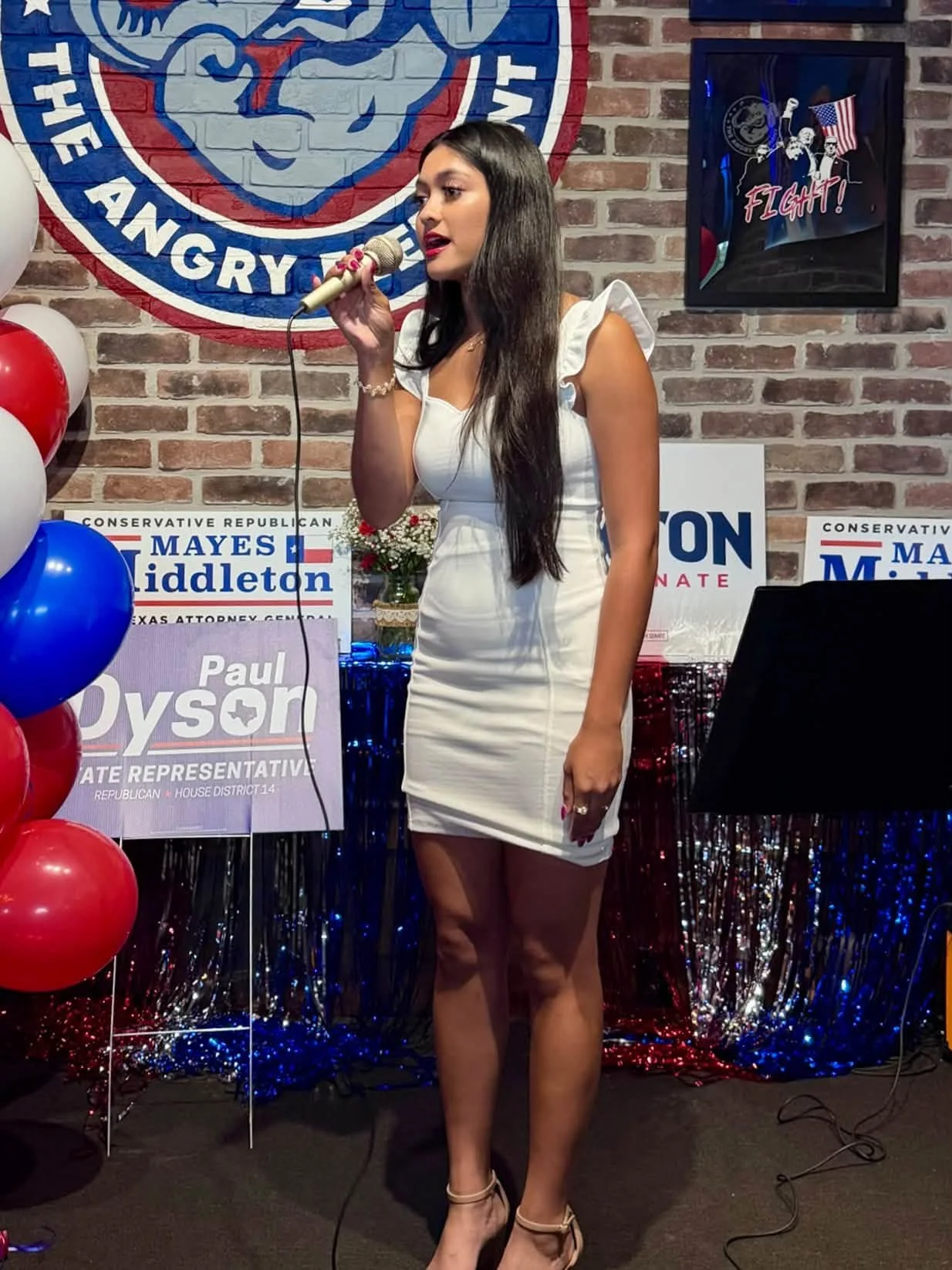 A woman with long dark hair standing on a decorated stage holding a microphone, wearing a white fitted dress with ruffled sleeves, in front of political campaign signs and patriotic decorations.