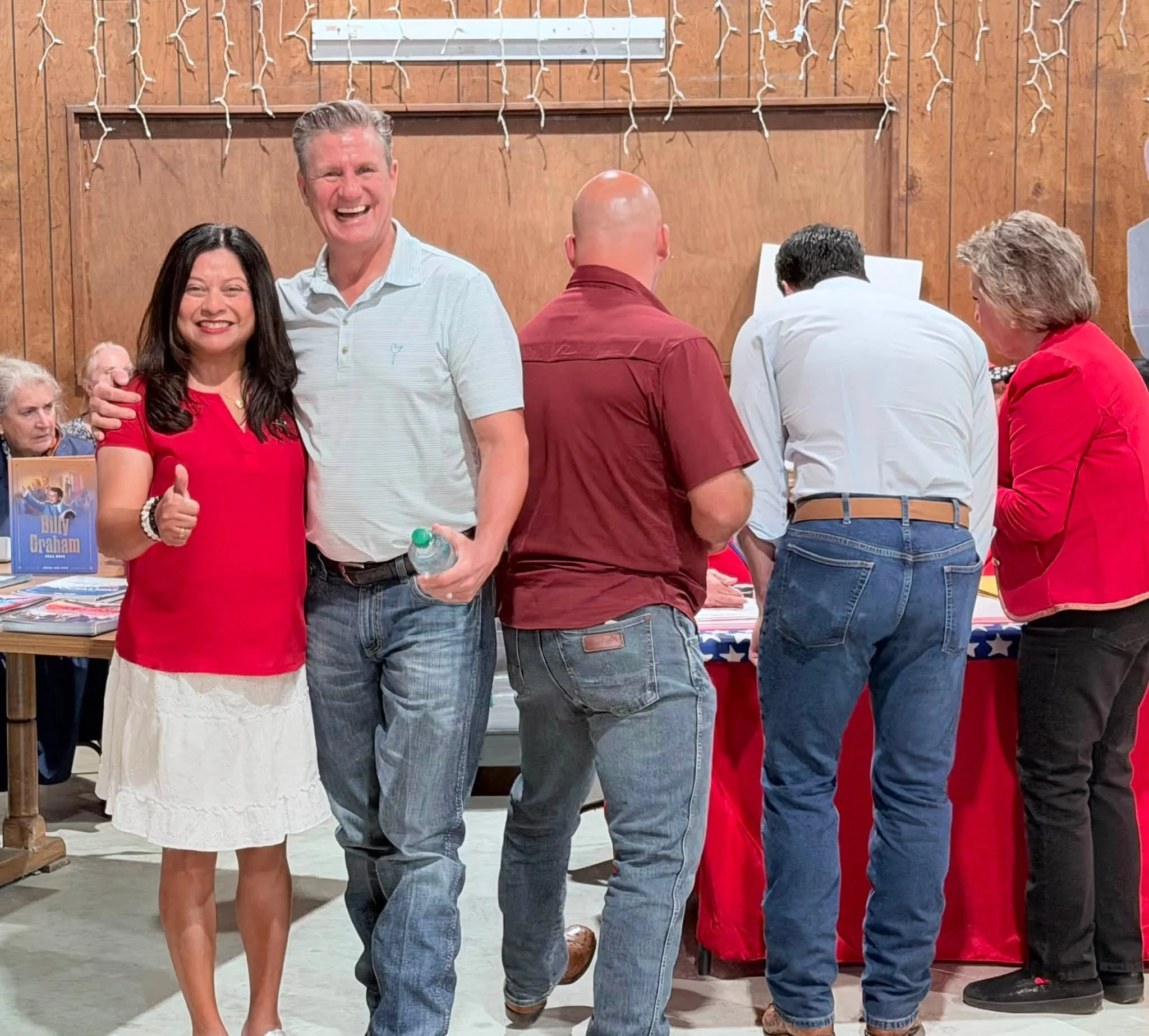 Two smiling people, a woman in a red top and a man in a light blue shirt, standing in front of a group of people in a room with wood paneling and holiday string lights.