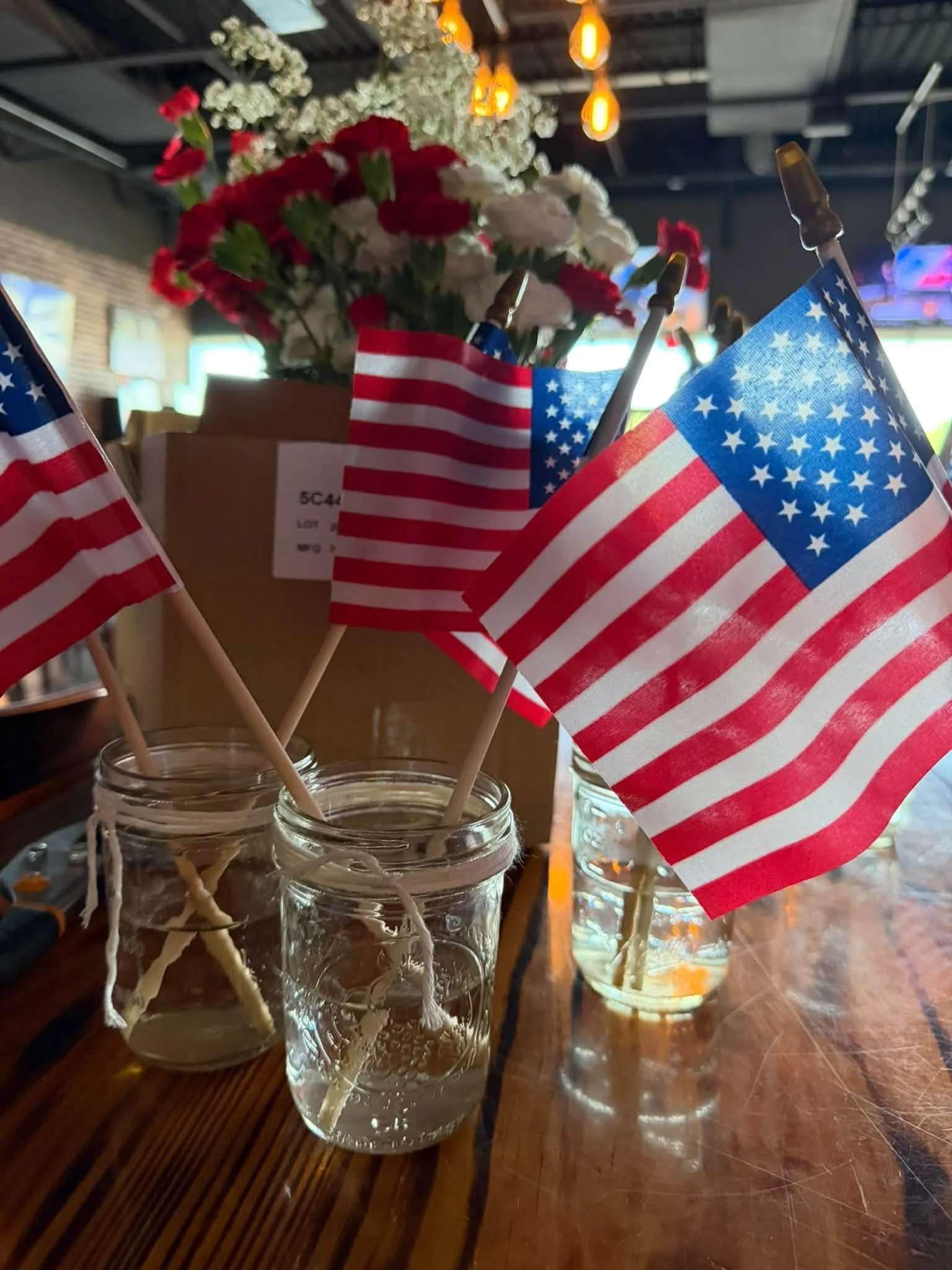 Multiple small American flags in glass jars on a wooden table with a floral arrangement in the background and warm hanging lights above.