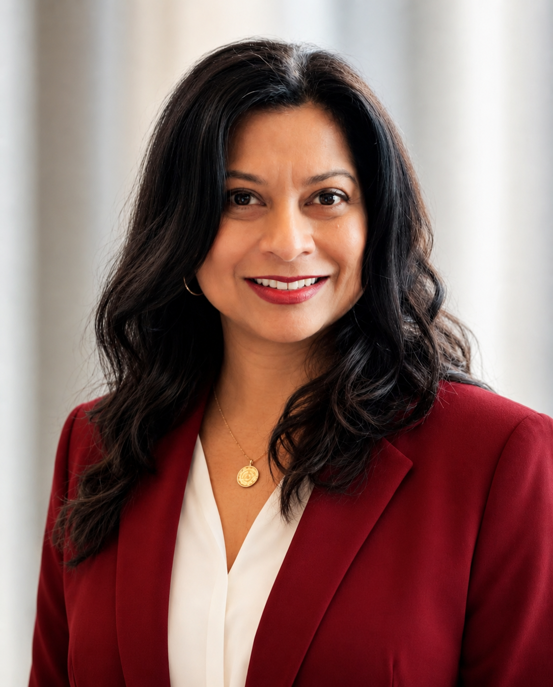 A woman with dark wavy hair, wearing a red blazer, white blouse, and gold jewelry, smiling in front of a neutral background.