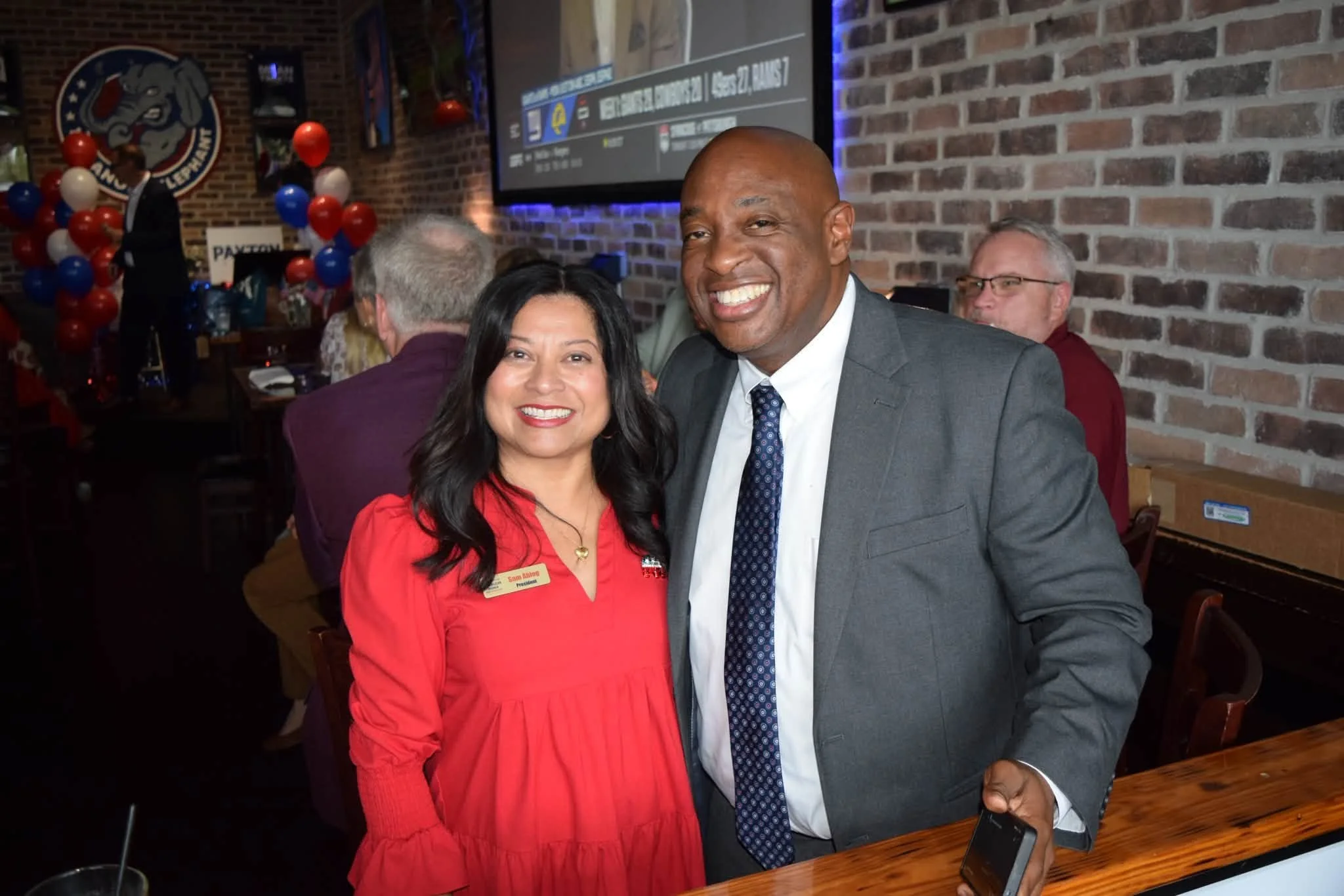 A smiling woman and a man in a gray suit with a tie posing together at an indoor event with brick walls, balloons, and a large screen in the background.