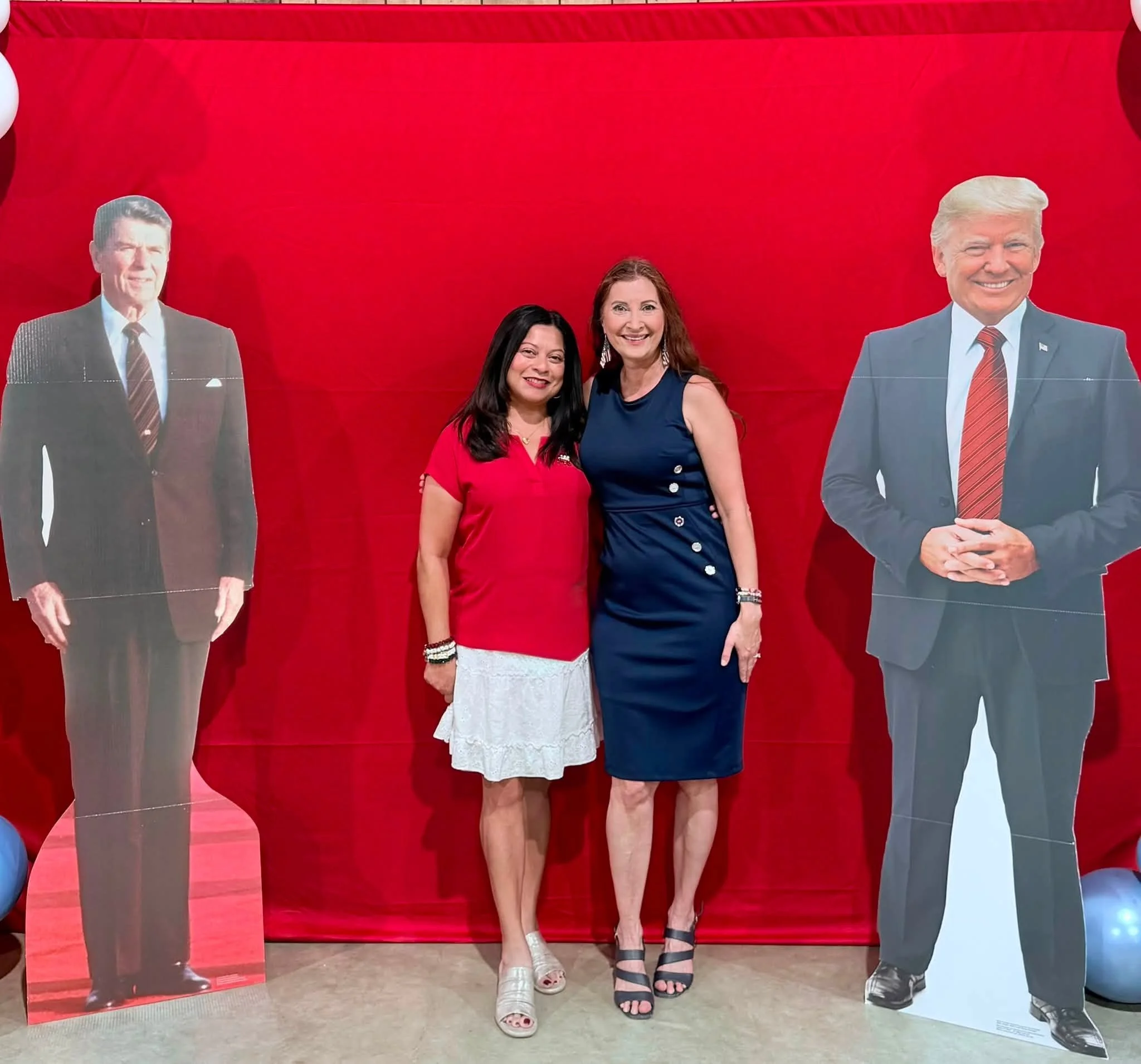 Two women smiling standing between life-sized cutouts of men in suits against a red background.