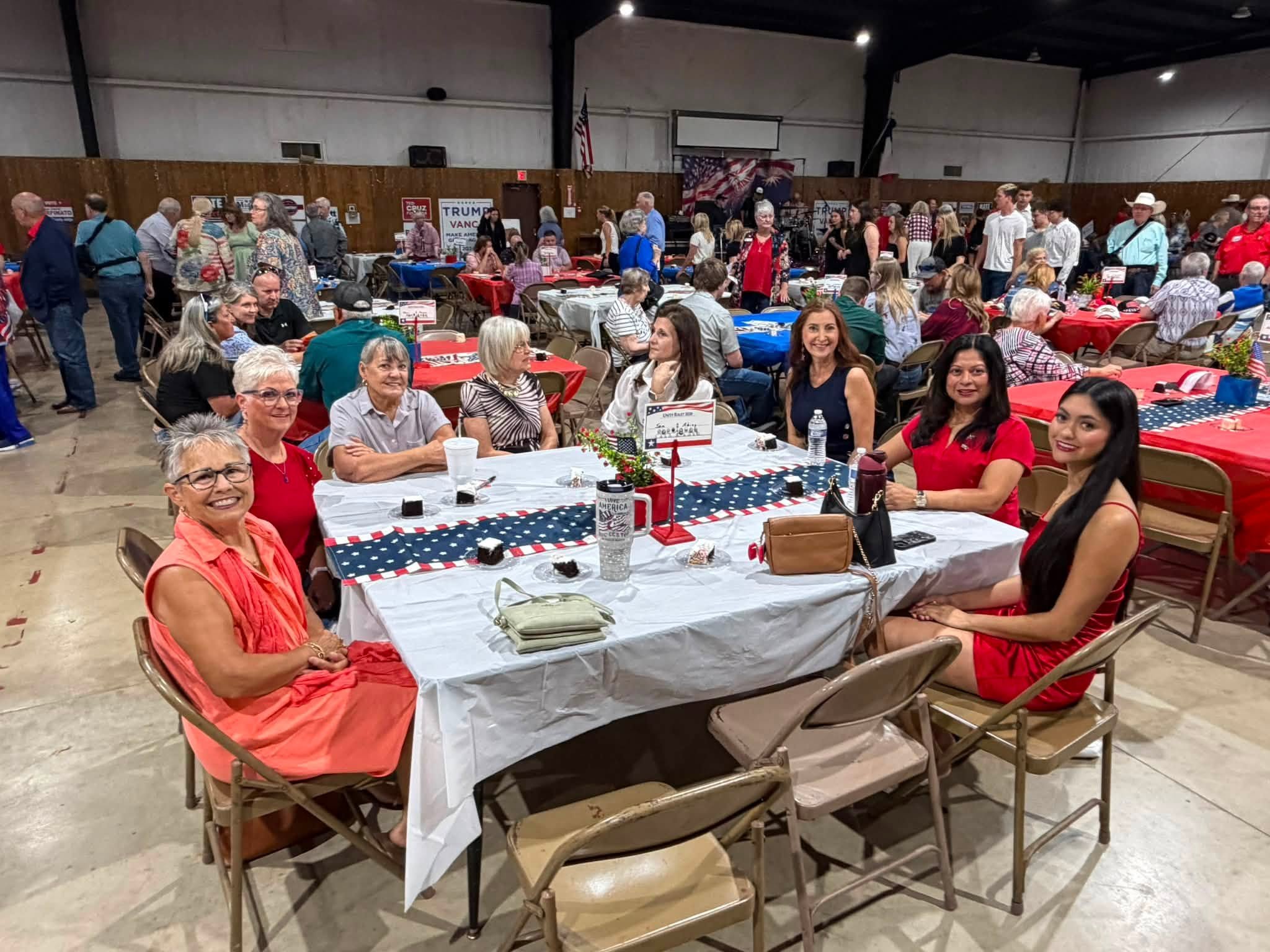 A large indoor event with numerous people seated at decorated tables with red, white, and blue patriotic themes. Many people are smiling and engaging with each other, with a backdrop of American flags and political campaign signs.