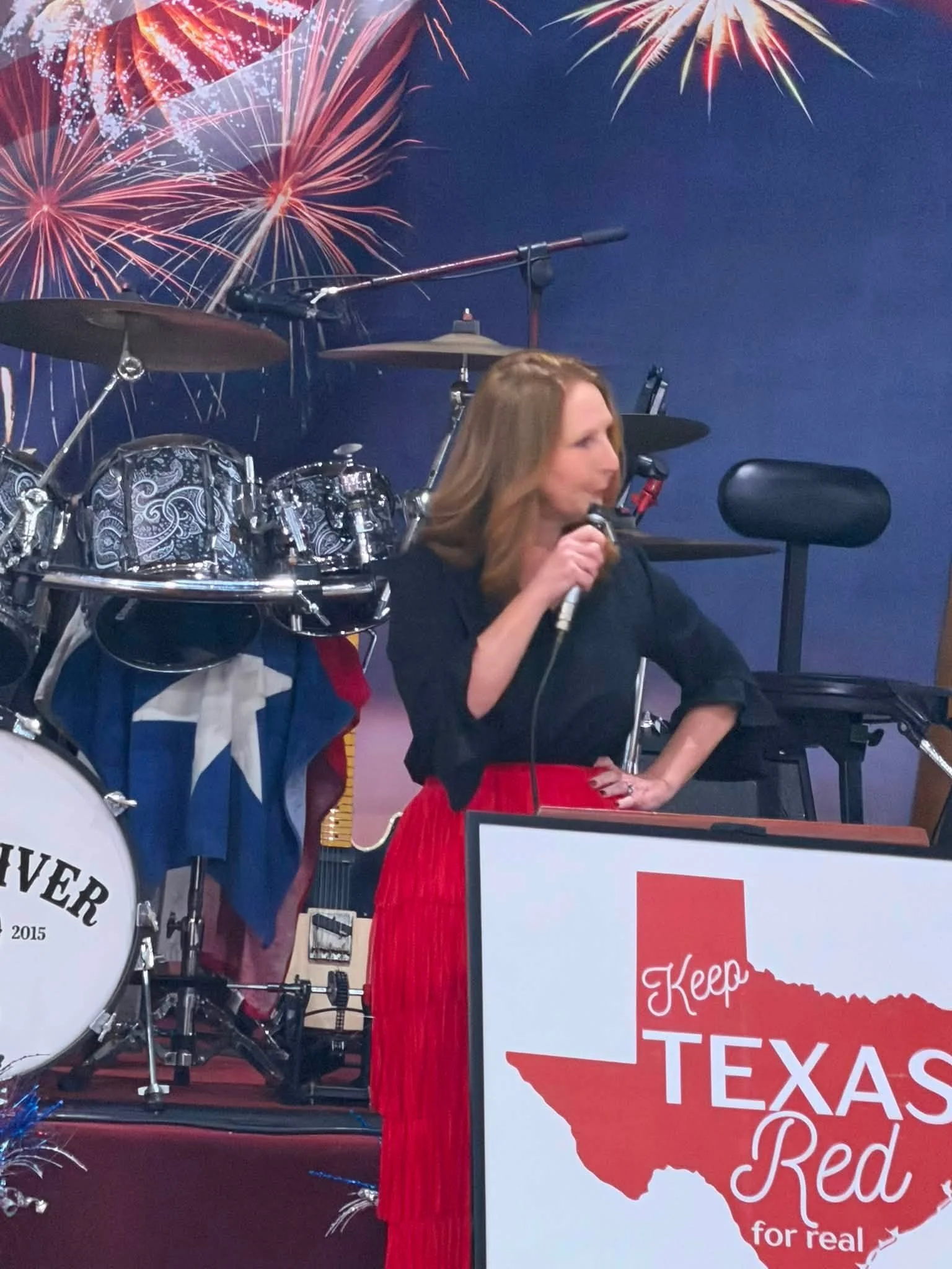 A woman singing into a microphone at a celebration event with fireworks display in the background, and a sign reading 'Keep Texas Red' with an outline of the state of Texas.