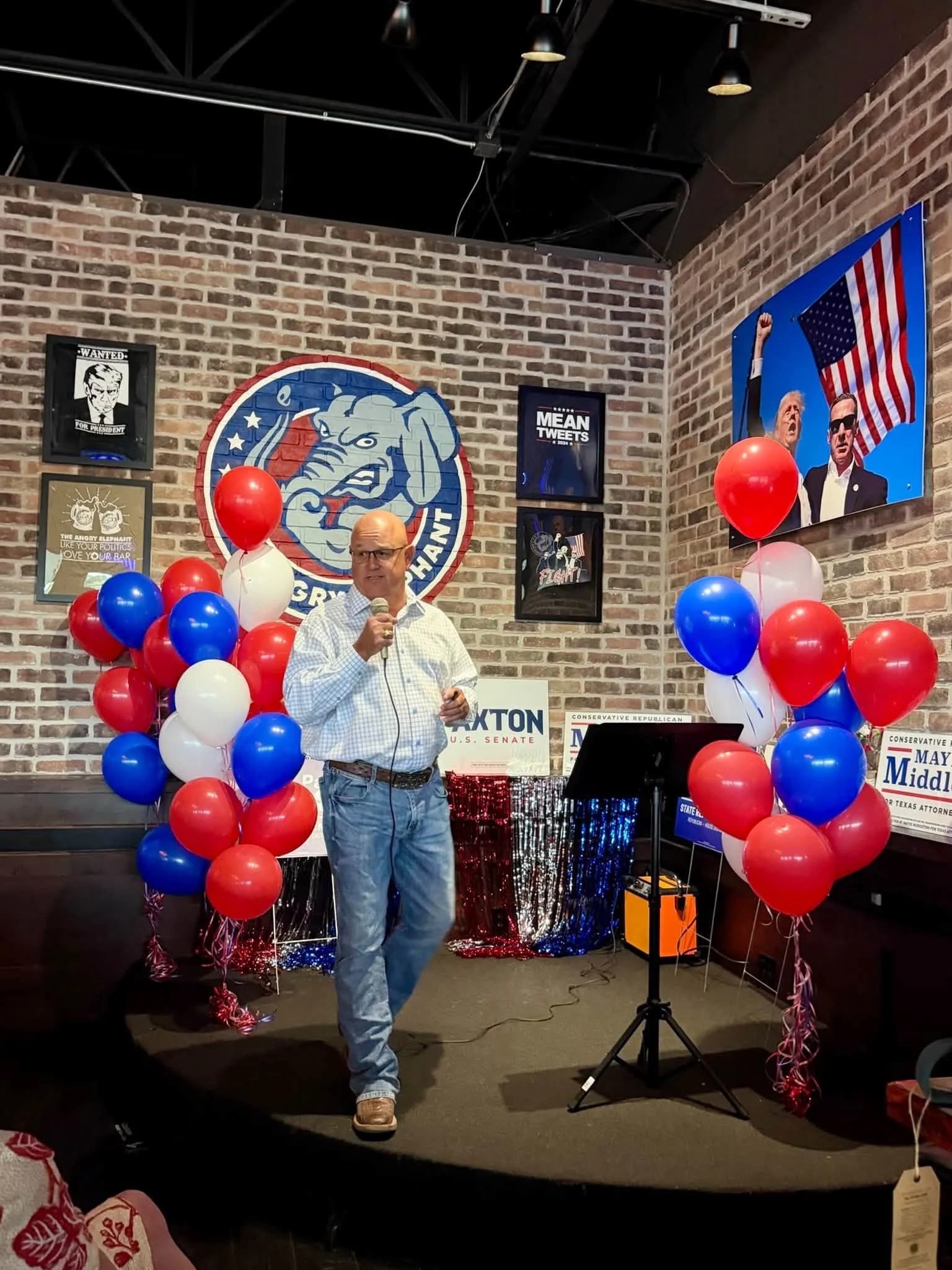 A man in a checkered shirt speaking into a microphone at a political event with red, white, and blue balloons and patriotic decorations, including a large flag mural and political posters on a brick wall.