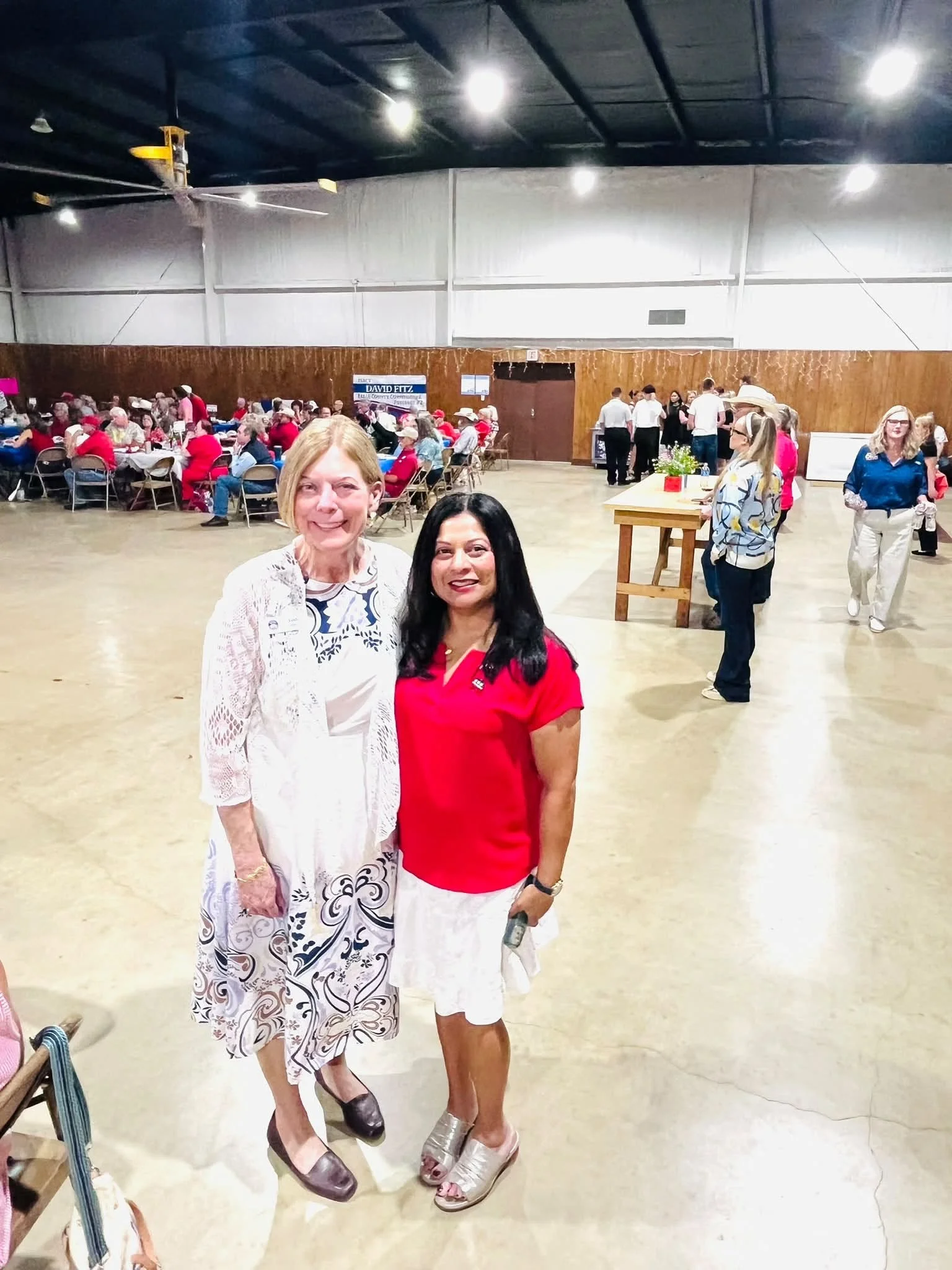 Two women standing together in a large indoor gathering, smiling at the camera. One woman is dressed in a white and black patterned dress and the other in a red top and white skirt. In the background, people are seated at tables, and some are standin