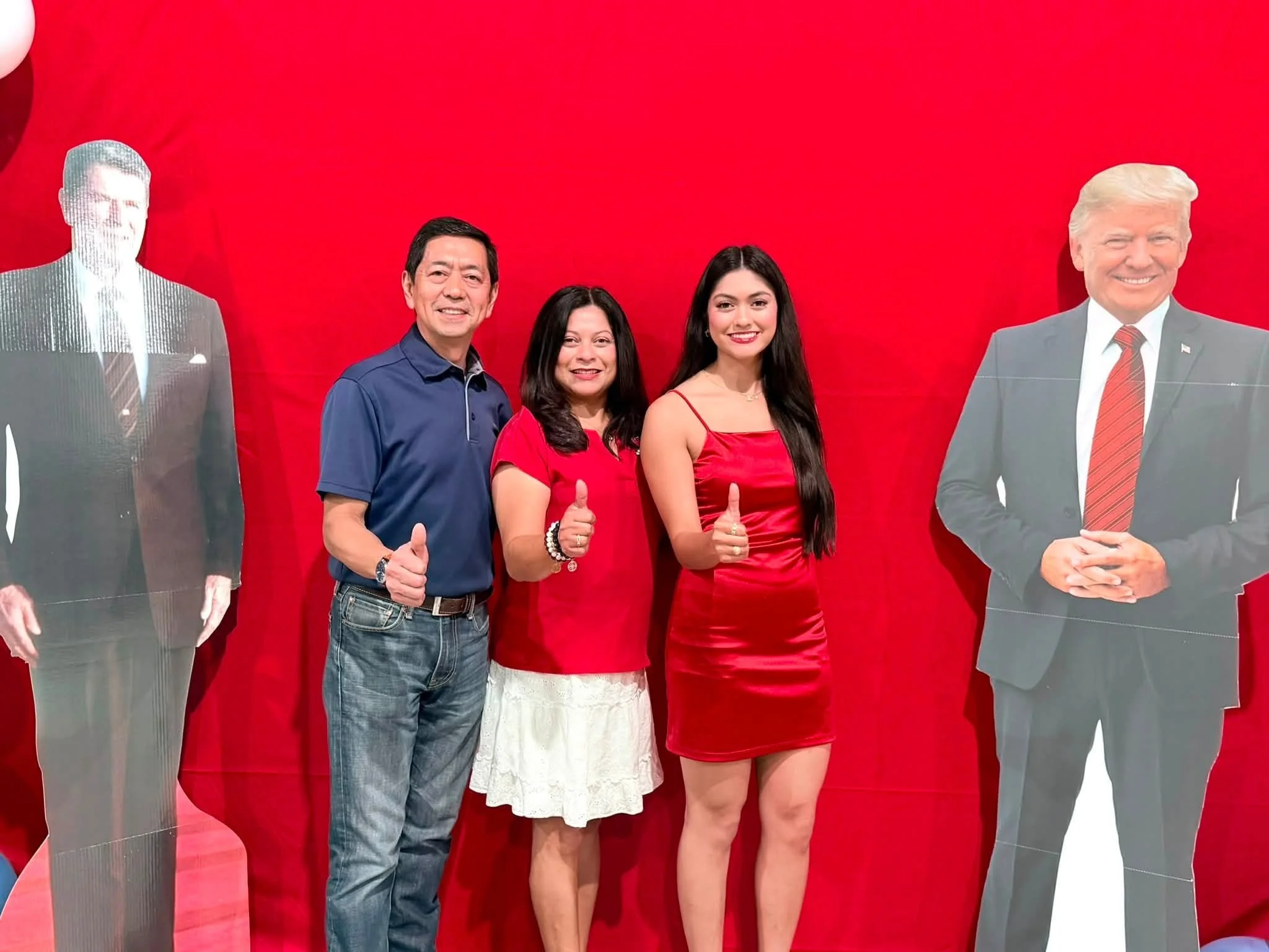 Four people standing in front of a red background, giving thumbs-up signs. Two cutout figures of men in suits are on the sides, with a man and woman in the center, smiling for the photo.